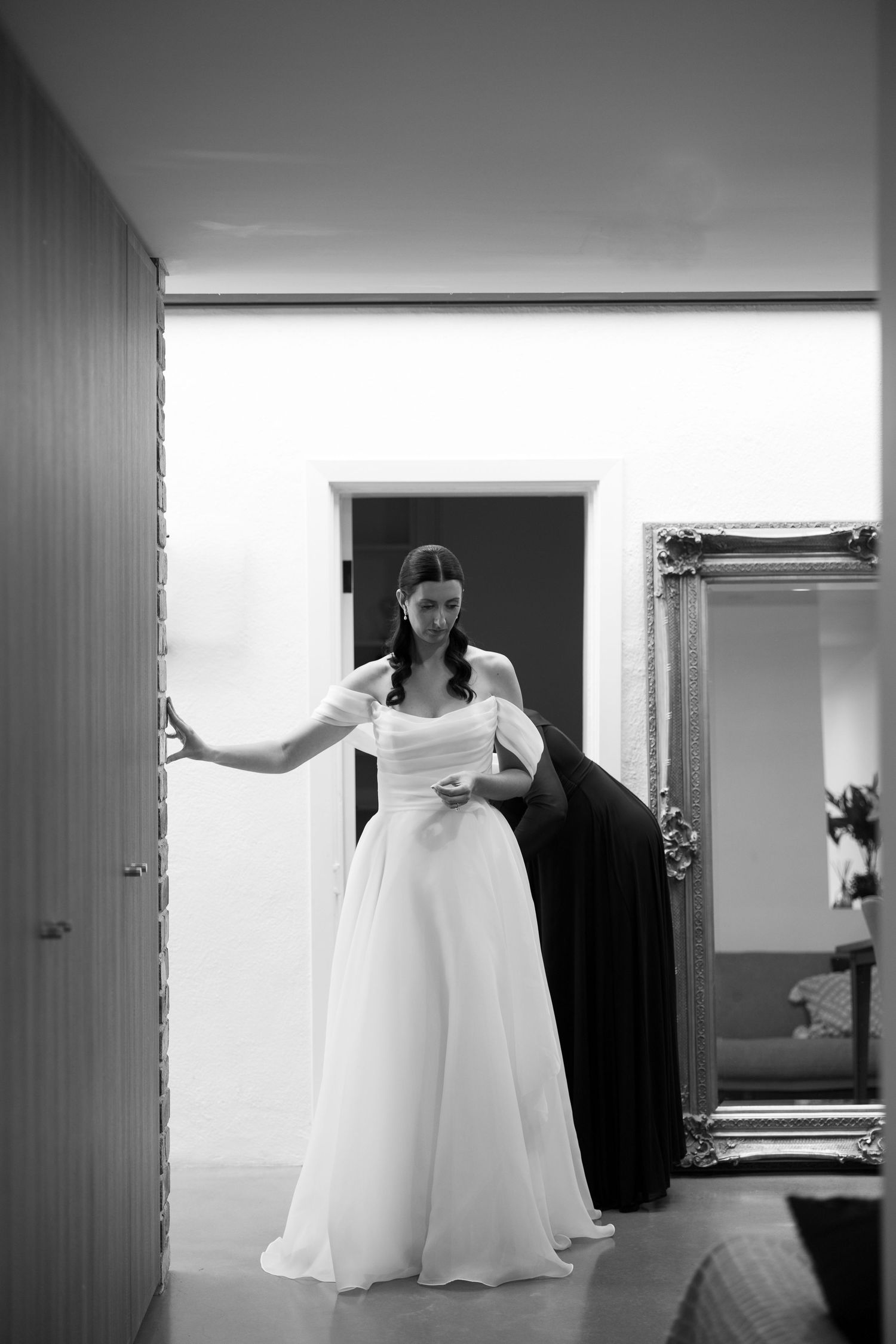Bride in white wedding gown holding dark jacket stands in doorway beside ornate mirror, black and white photo.