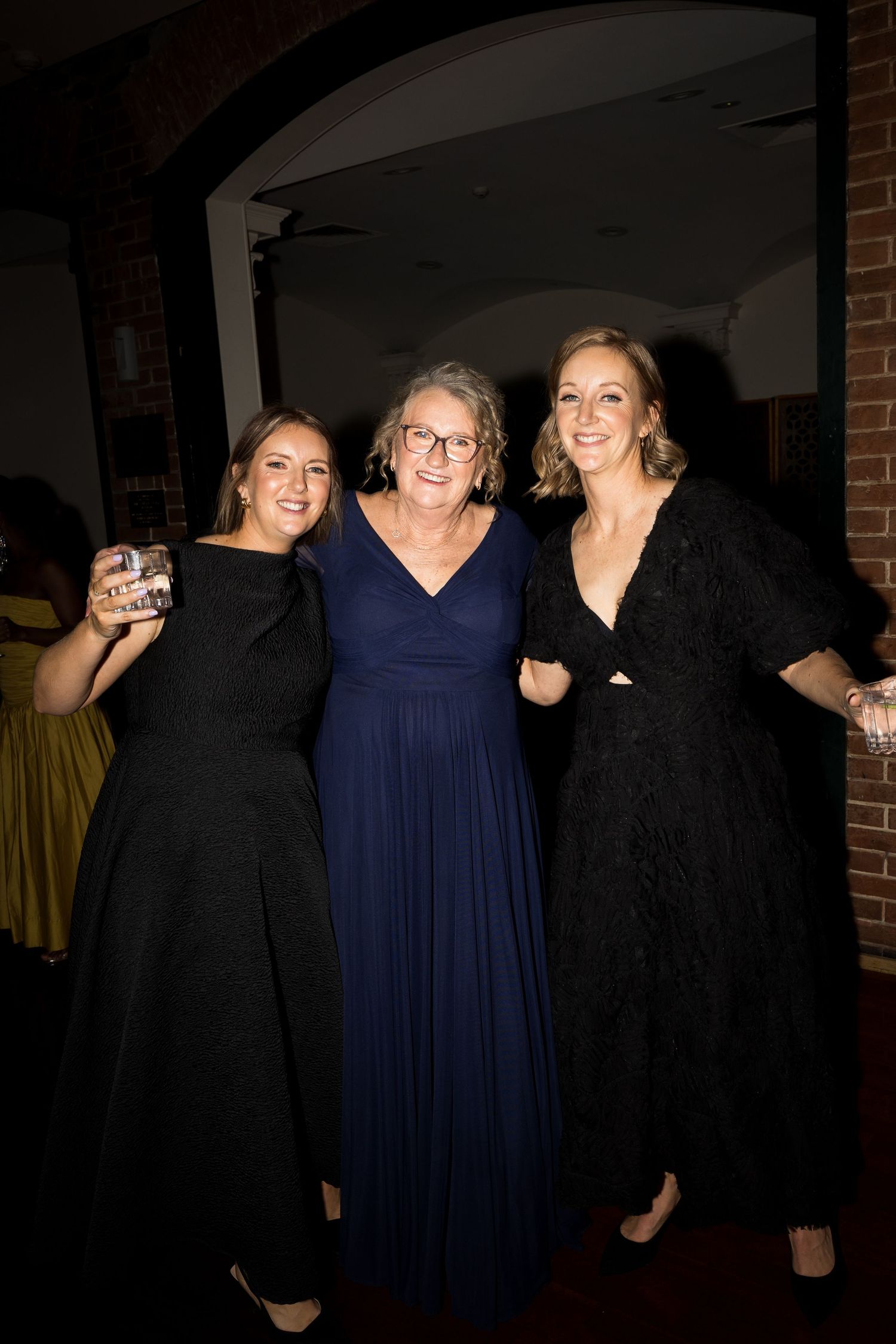 Three women in elegant black and navy gowns smiling together at a formal event.