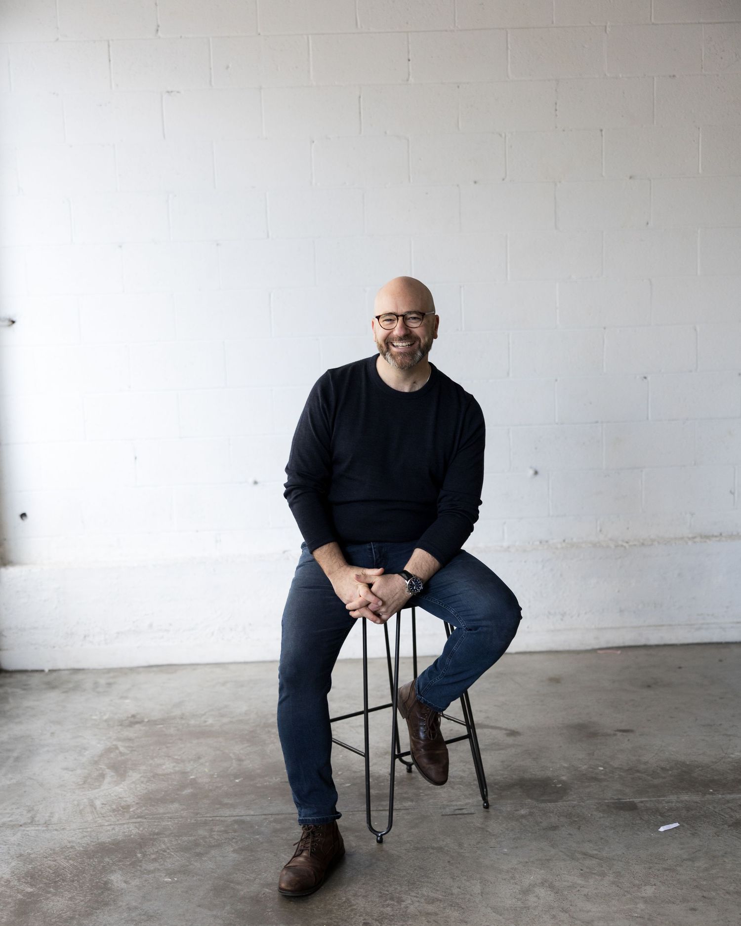 Bald man with glasses in black sweater and jeans sits smiling on a stool against a white brick wall.