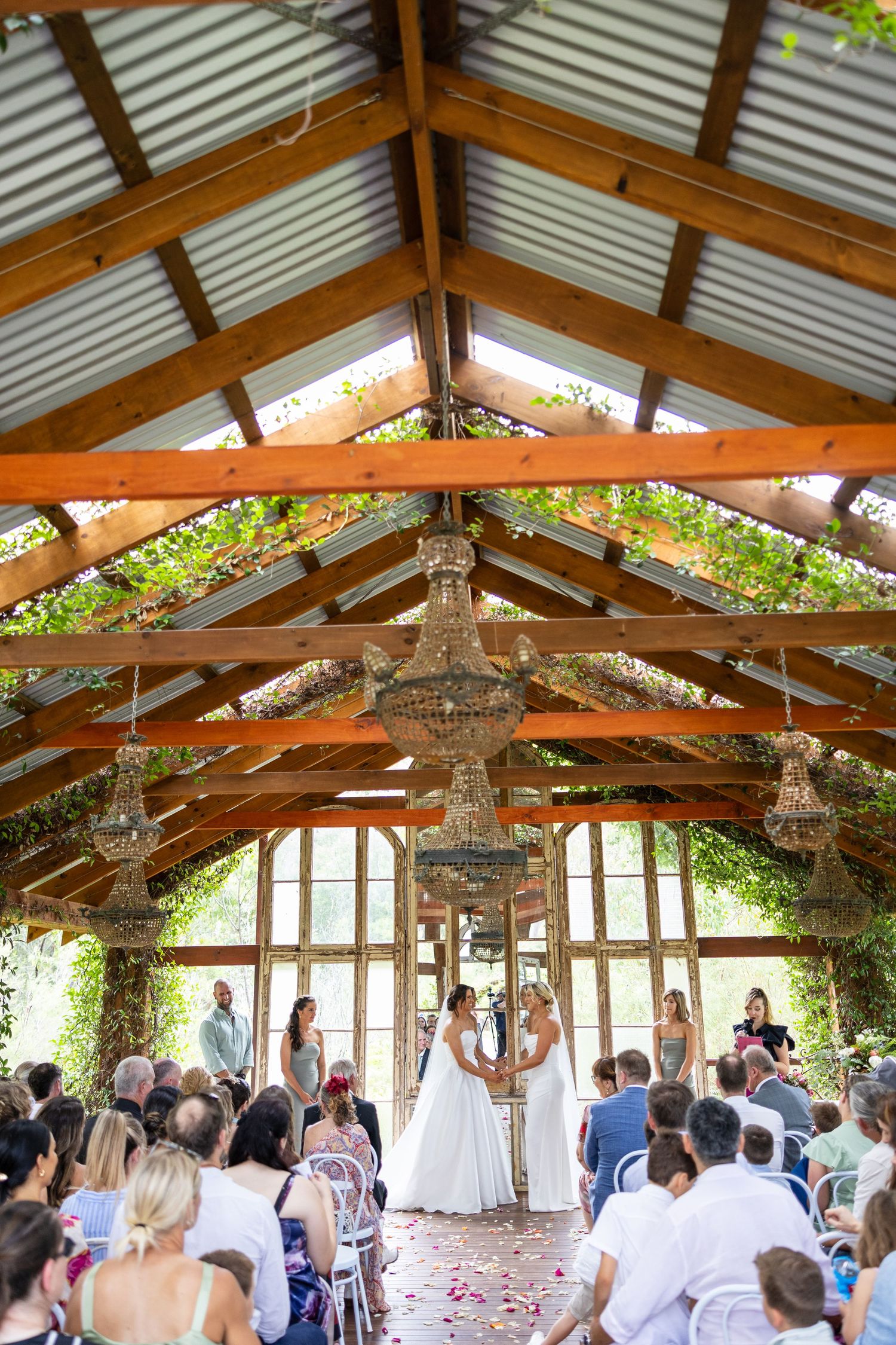 Two brides exchange vows beneath a grand chandelier in a stunning rustic timber-beamed wedding pavilion.