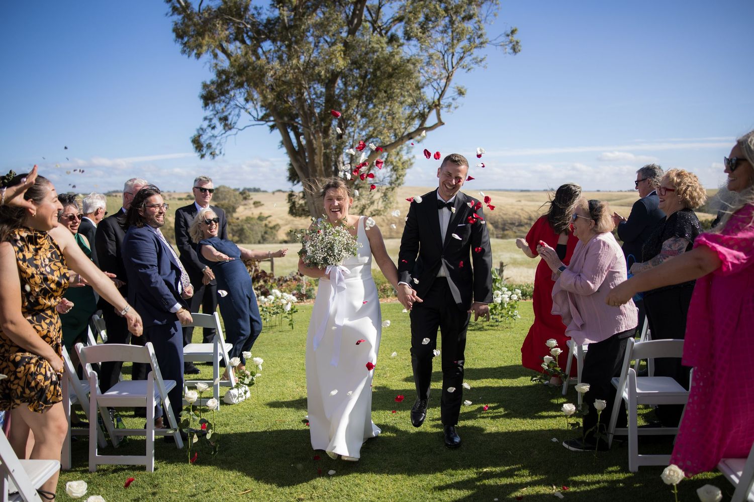 Bride and groom walk down outdoor wedding aisle as guests toss flower petals under blue sky.