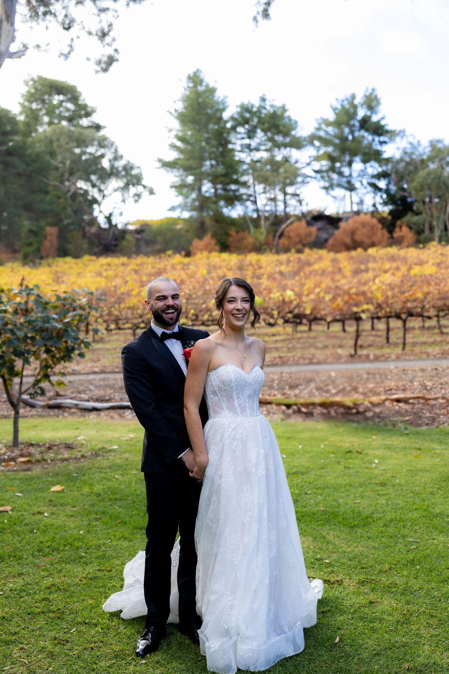 Smiling couple in wedding attire hold hands on green lawn with golden autumn vineyard rows in the background.