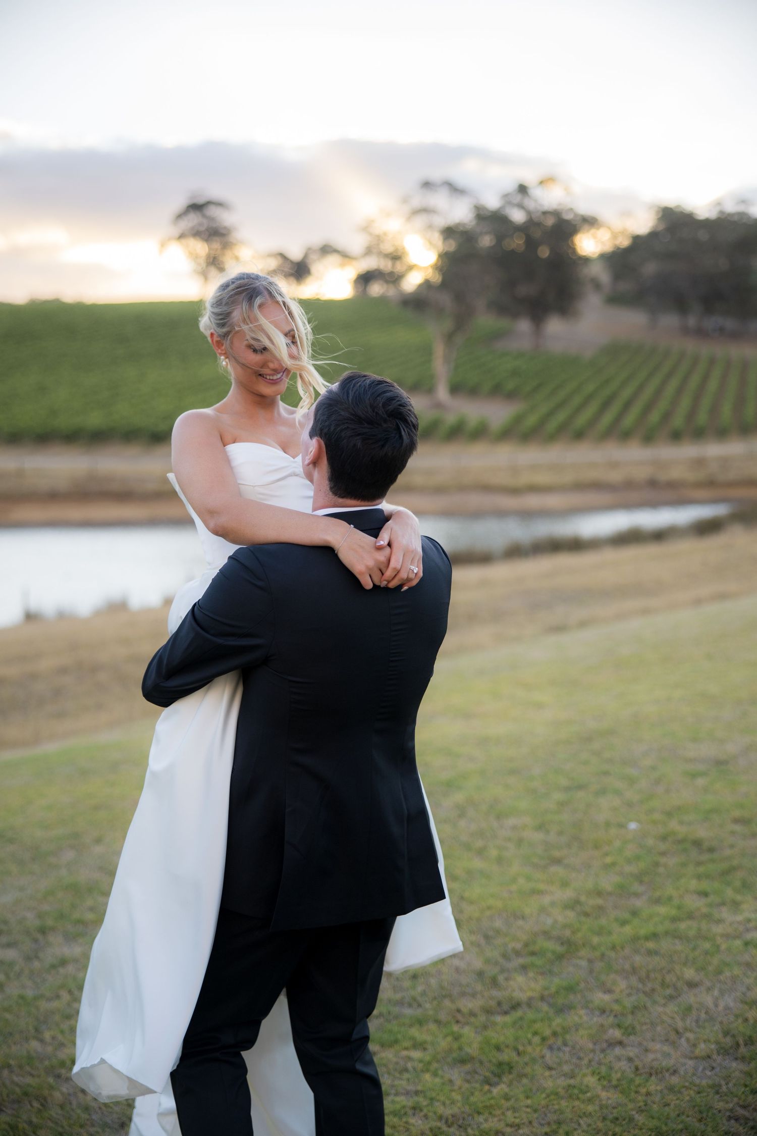 Groom lifts smiling bride in strapless white dress near vineyard pond at sunset.