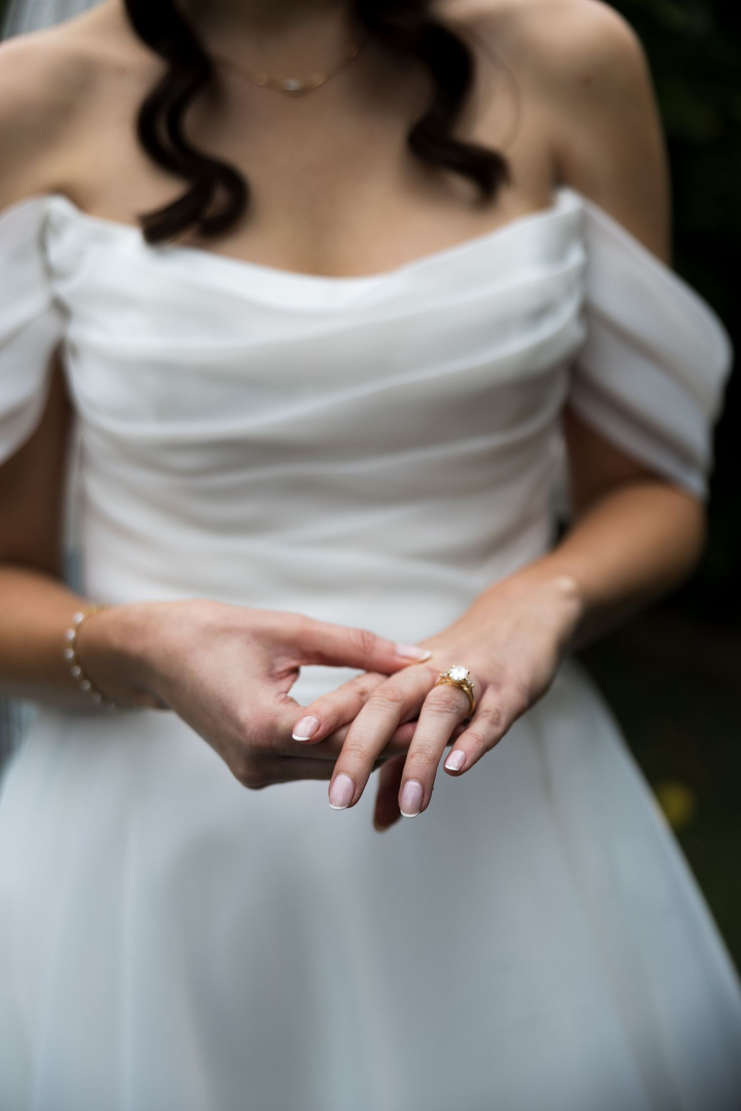 Bride in white off-shoulder gown showcasing elegant gold engagement ring and bracelet with French manicure.