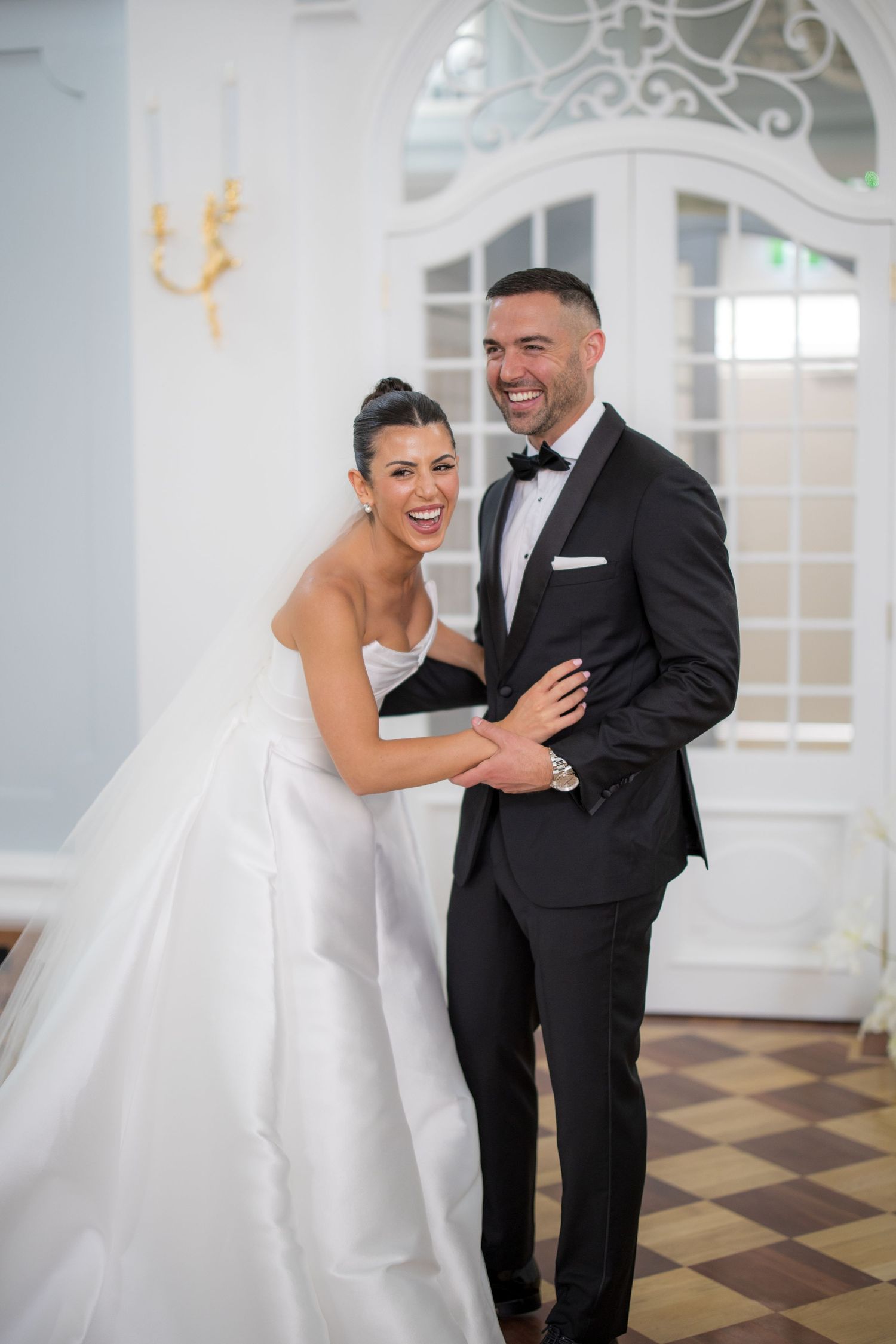 Laughing bride and groom embrace joyfully in bright elegant ballroom, she in white ballgown, he in tuxedo.