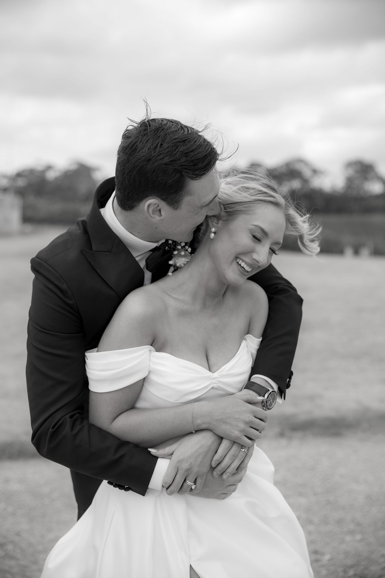 Black and white photo of groom embracing laughing bride from behind at outdoor wedding.