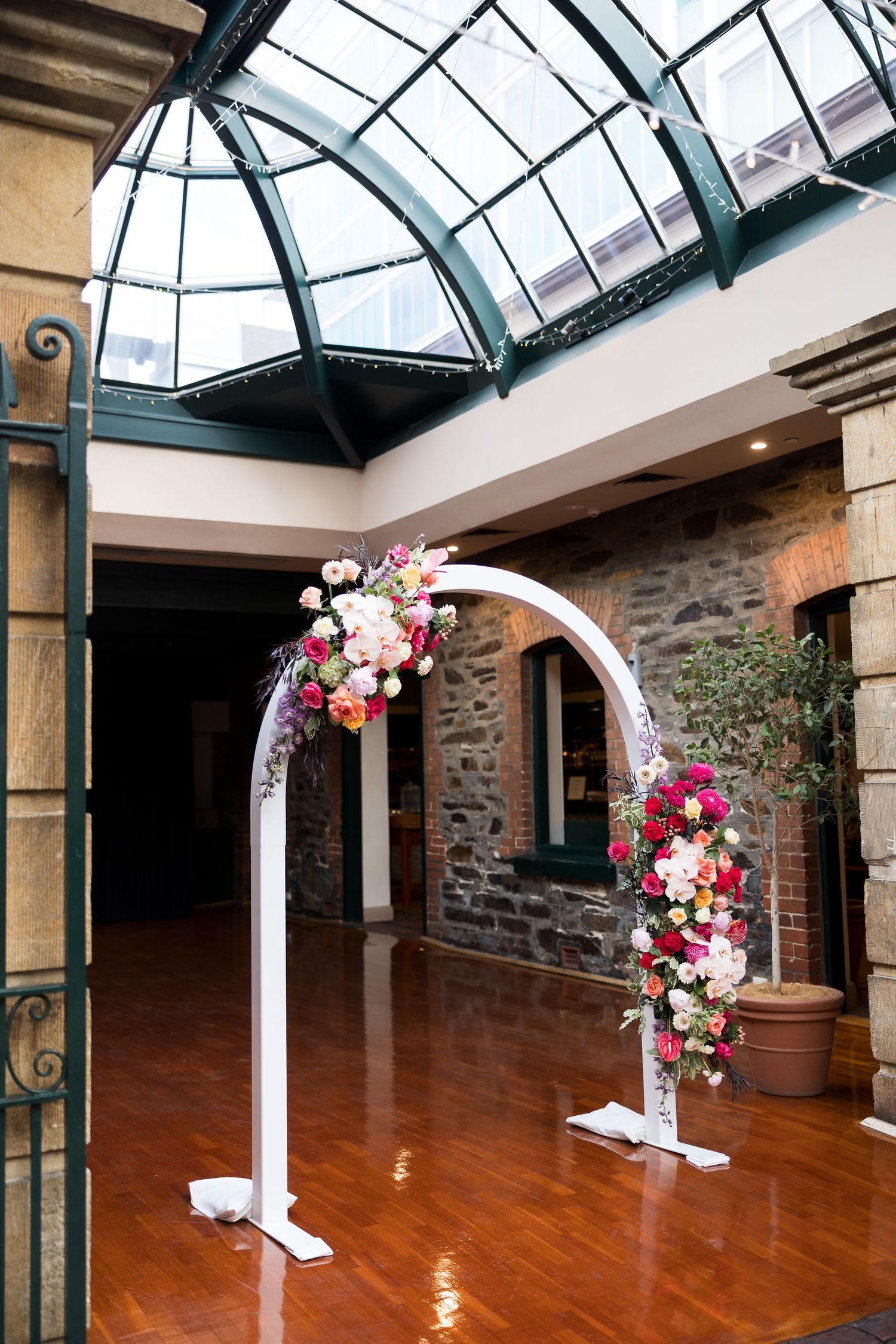 White floral wedding arch with pink and red blooms in elegant stone venue with glass skylight ceiling.
