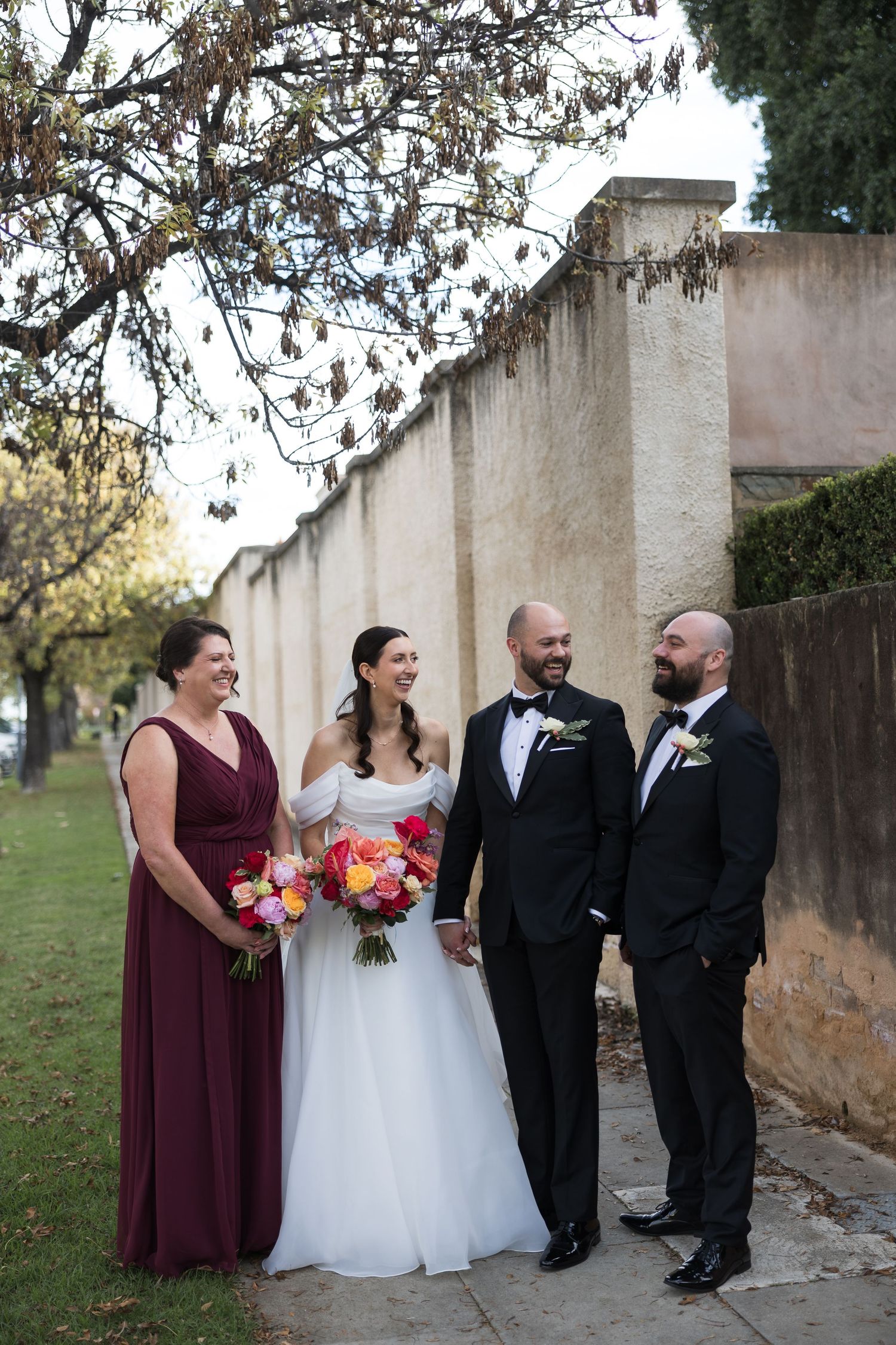 Wedding party of four posing outdoors, bride in white gown with bouquet, bridesmaid in burgundy, grooms in black tuxedos.