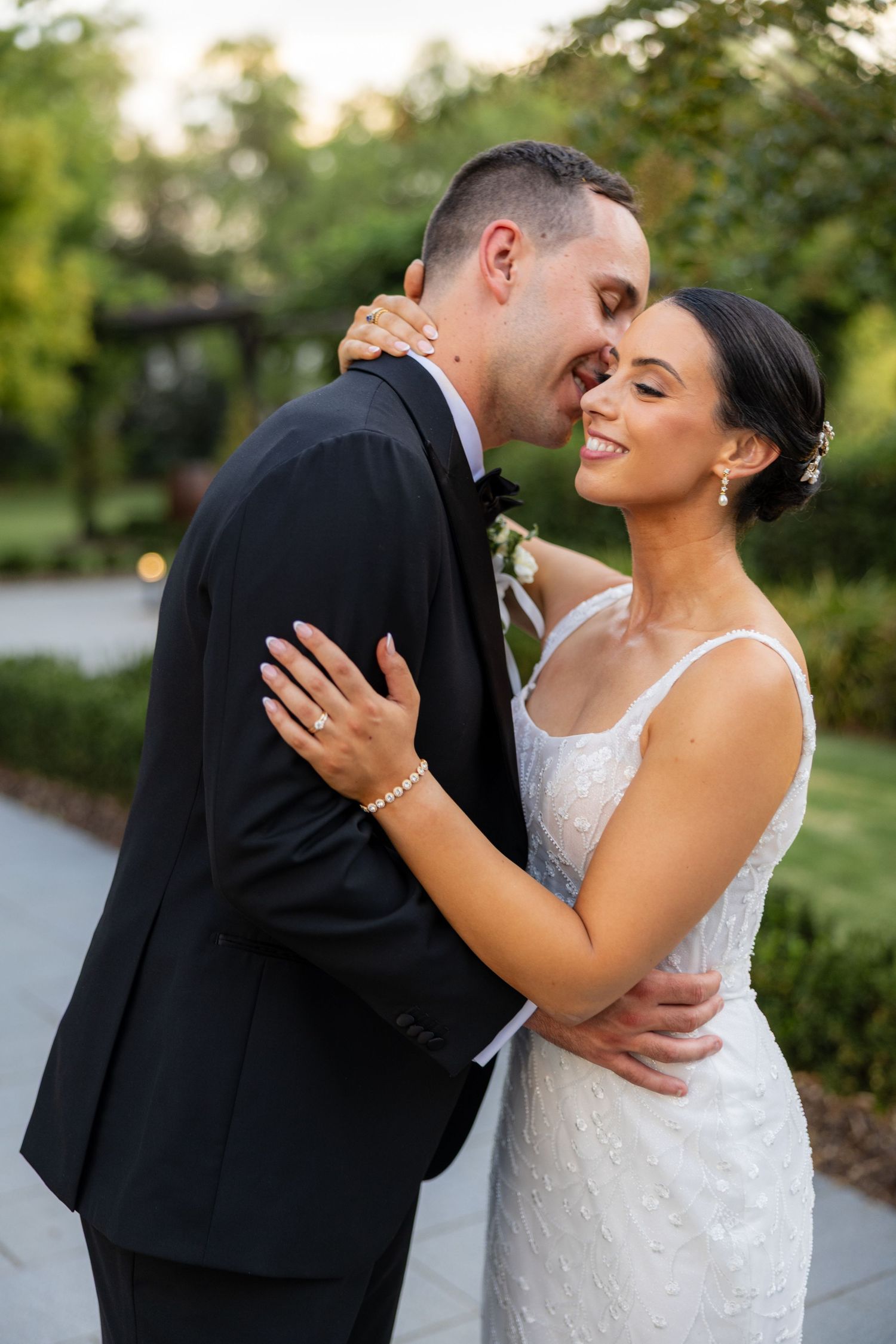 Groom in black tuxedo tenderly embracing smiling bride in beaded gown in a garden at dusk.