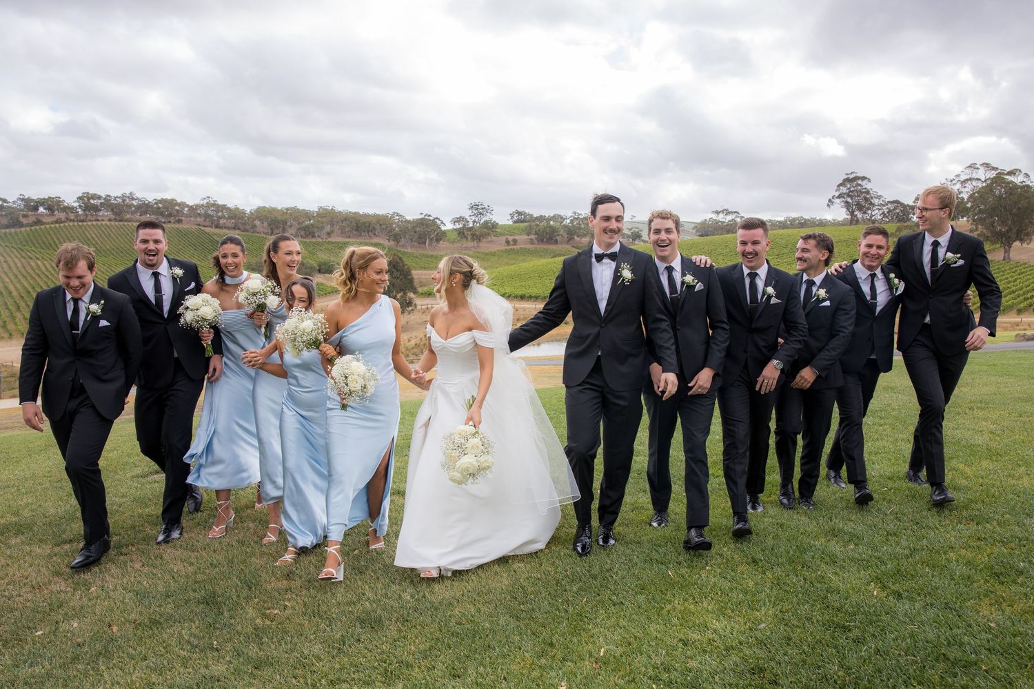 Wedding party with bridesmaids in blue dresses and groomsmen in black suits pose at vineyard.