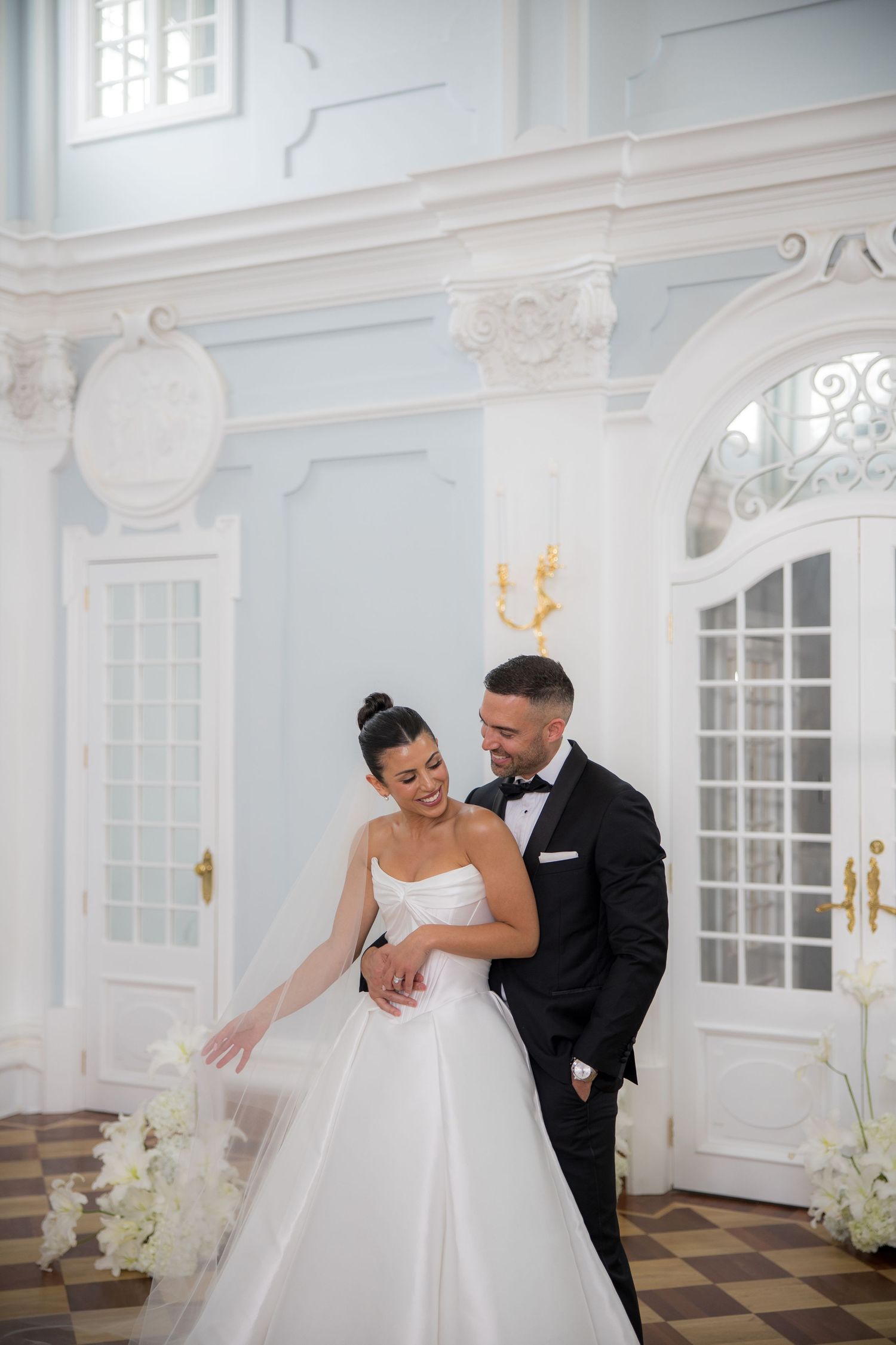 Bride and groom share joyful moment in elegant blue ballroom, admiring her white ballgown wedding dress.