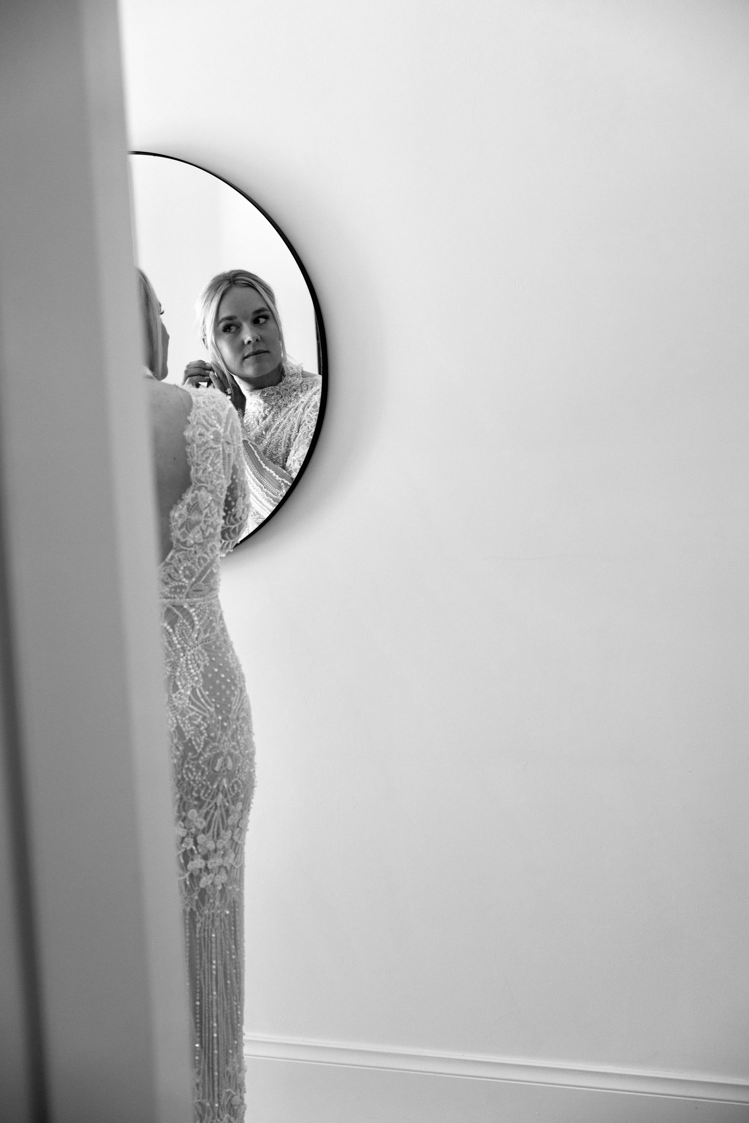 Black and white portrait of a bride in a beaded lace gown reflected in a round mirror against a white wall.