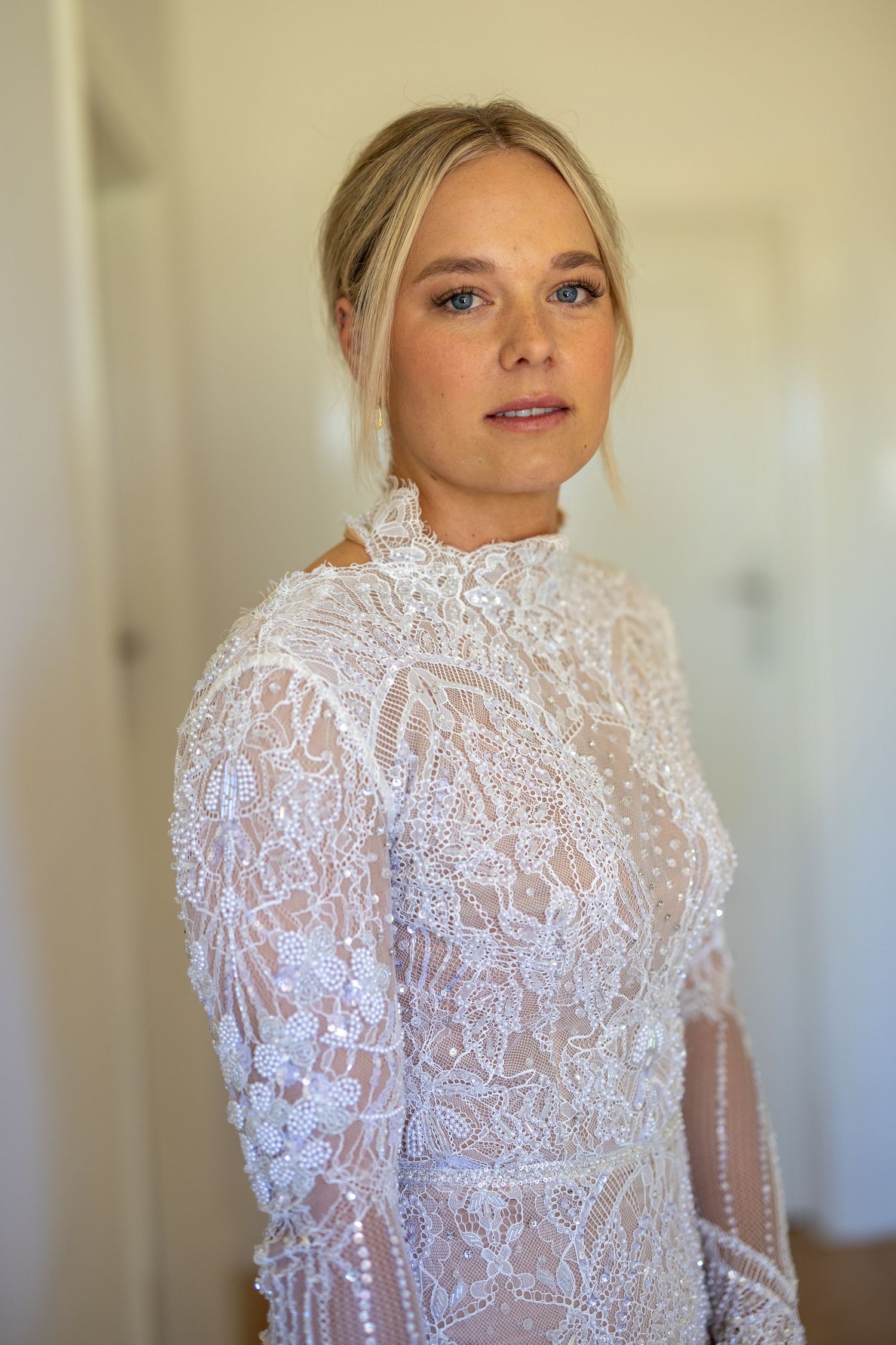 Portrait of a blonde bride in an intricate beaded high-neck lace wedding gown indoors.