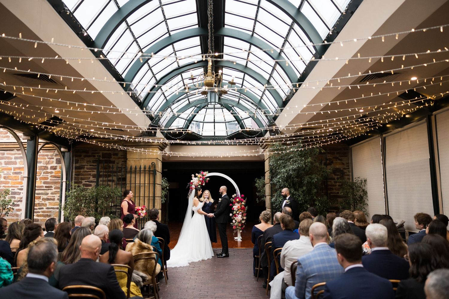 Wedding ceremony in elegant glass-roofed venue with string lights, floral arch, and seated guests.