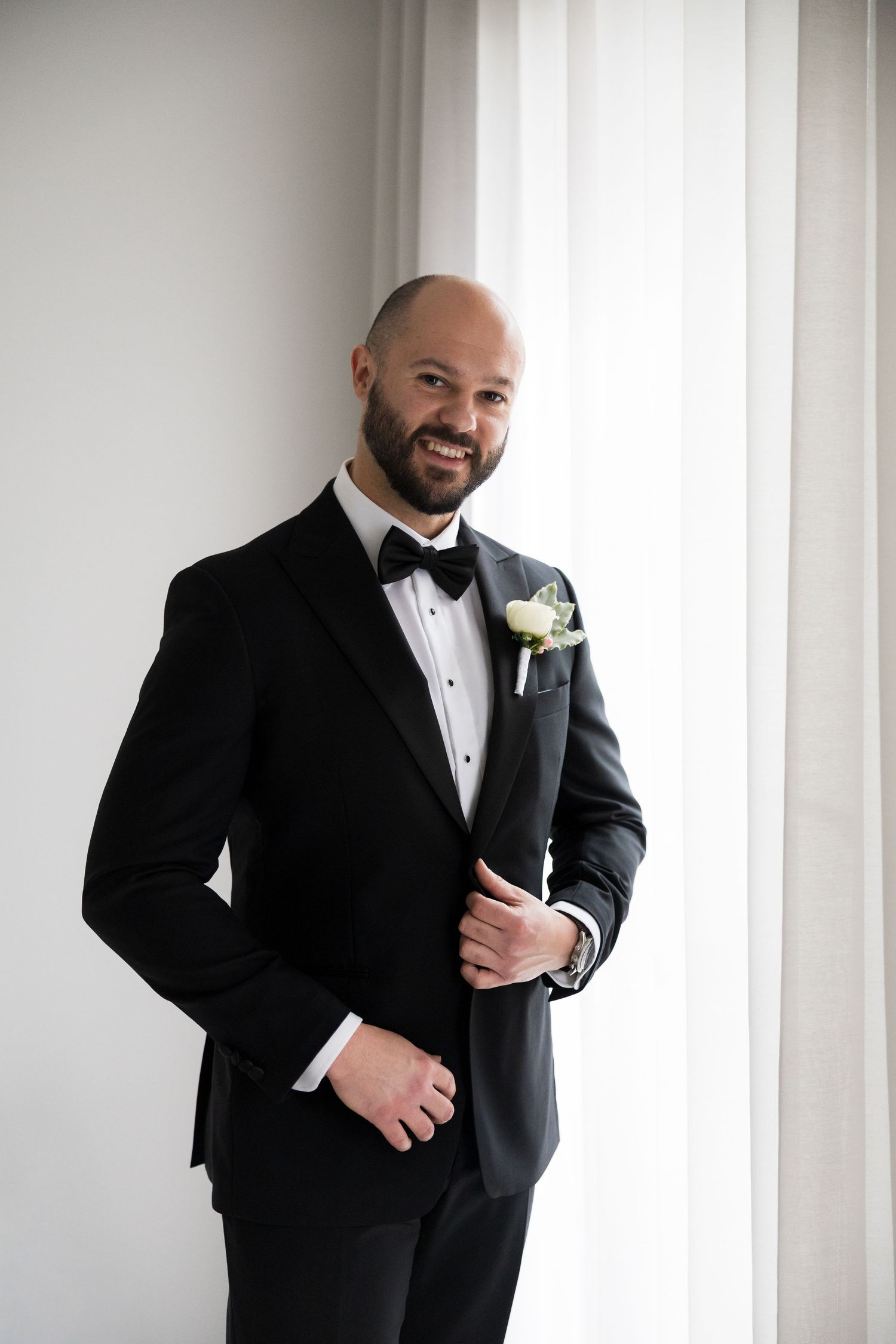 Smiling groom in black tuxedo with bow tie and white boutonniere stands confidently near a bright window.