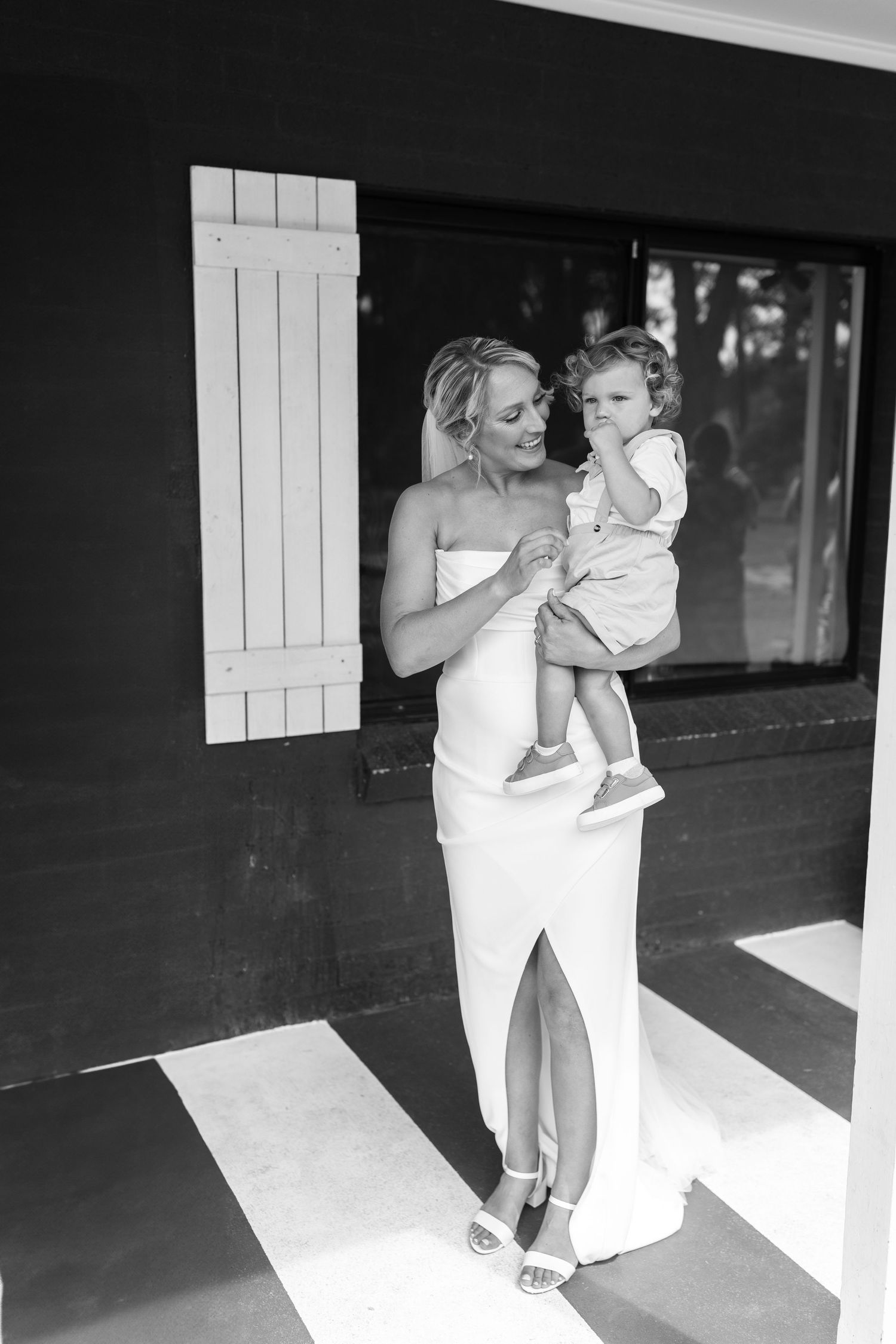 Black and white photo of bride in white gown holding a young toddler outside a rustic venue.