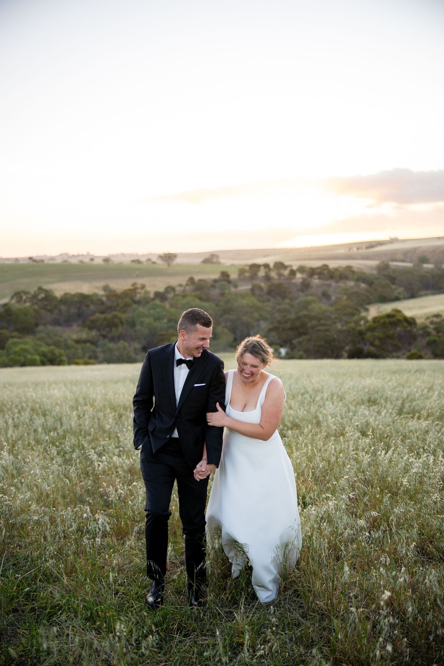 Bride and groom smiling together in a lush green field with rolling hills at sunset.