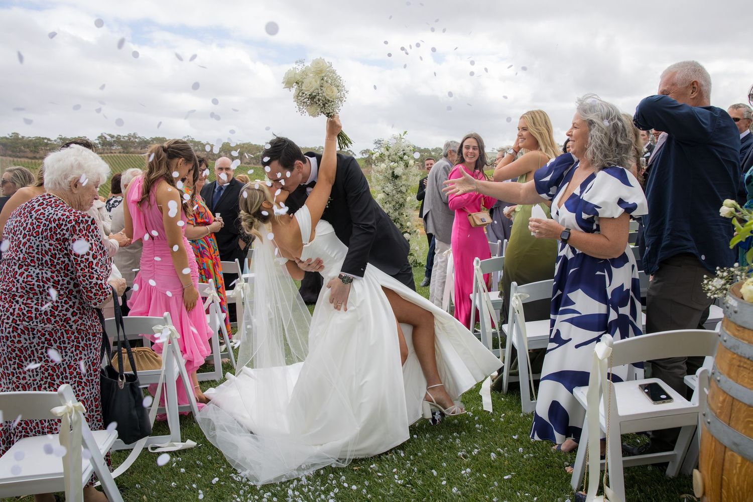 Groom dips bride for romantic kiss during confetti celebration as wedding guests cheer outdoors.