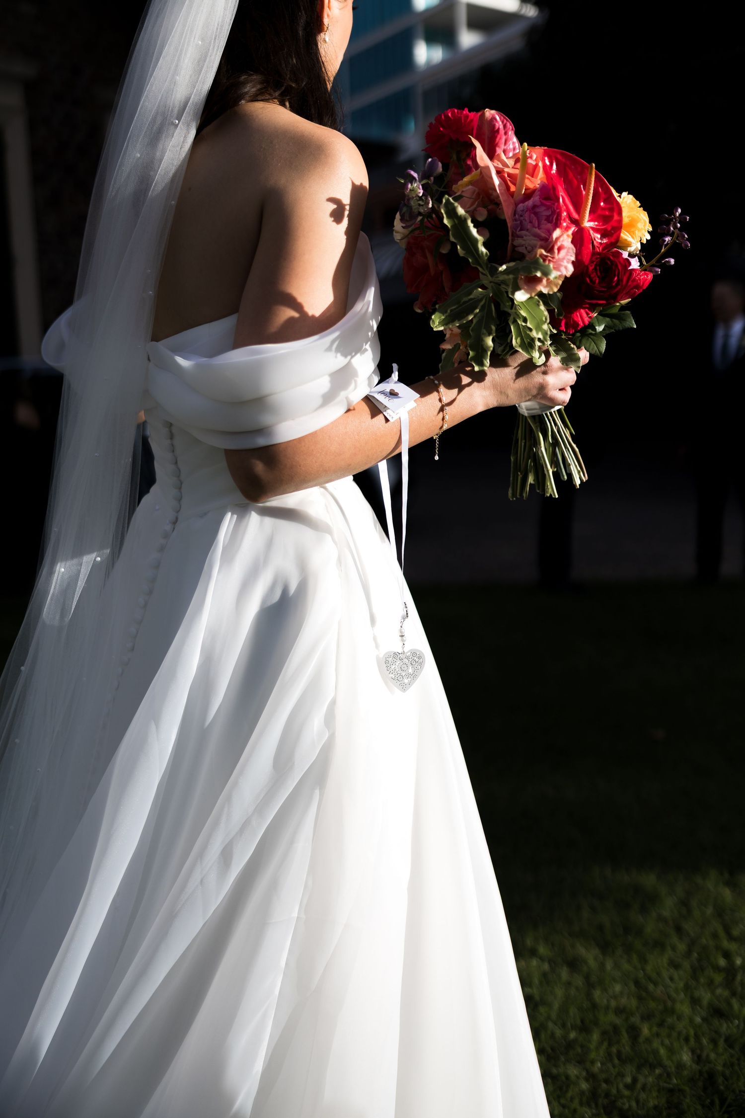 Bride in white off-shoulder gown holding vibrant pink and red floral bouquet, standing outdoors in dramatic lighting.
