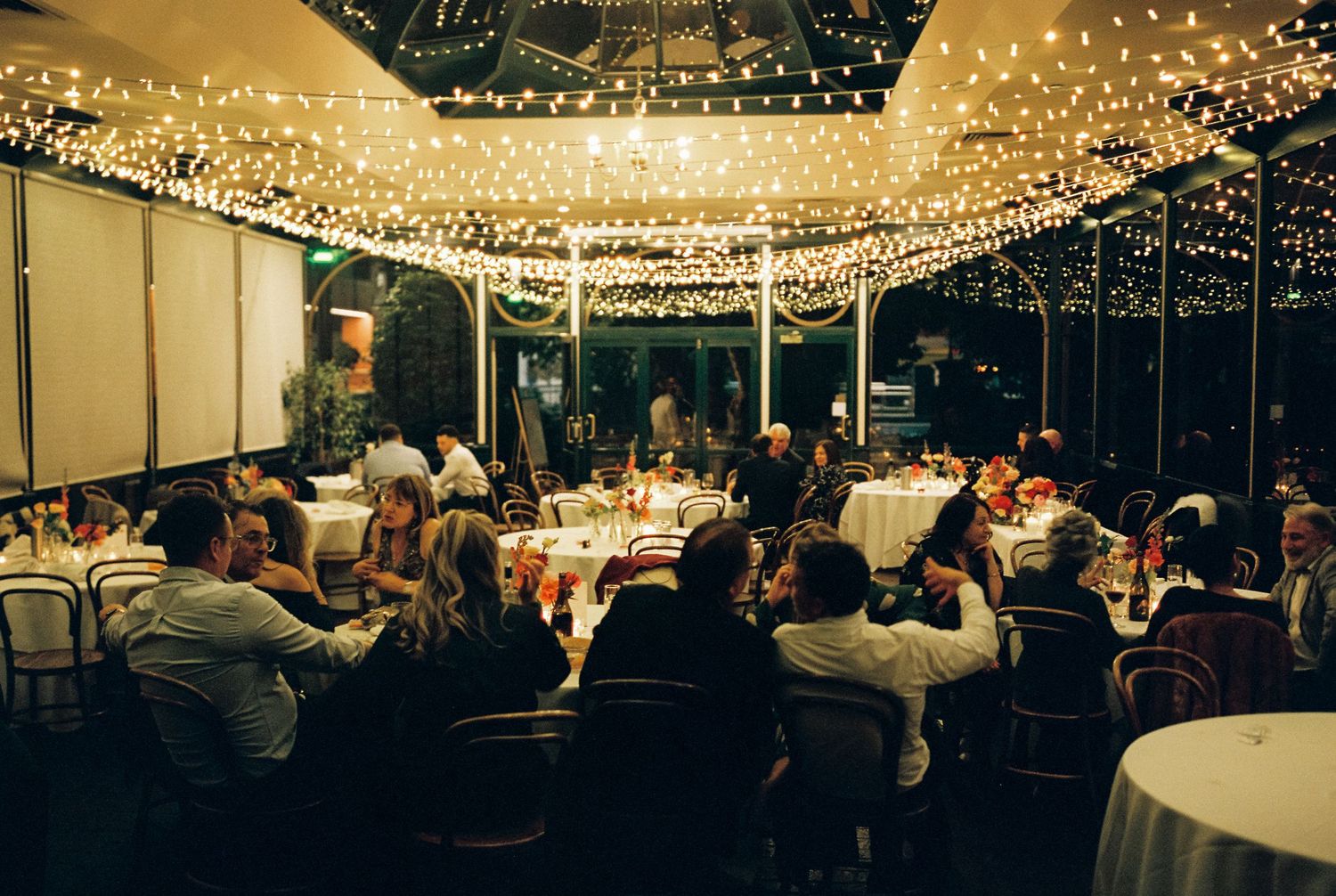 Elegant indoor wedding reception with fairy lights canopy, guests dining at round tables in a warmly lit venue.