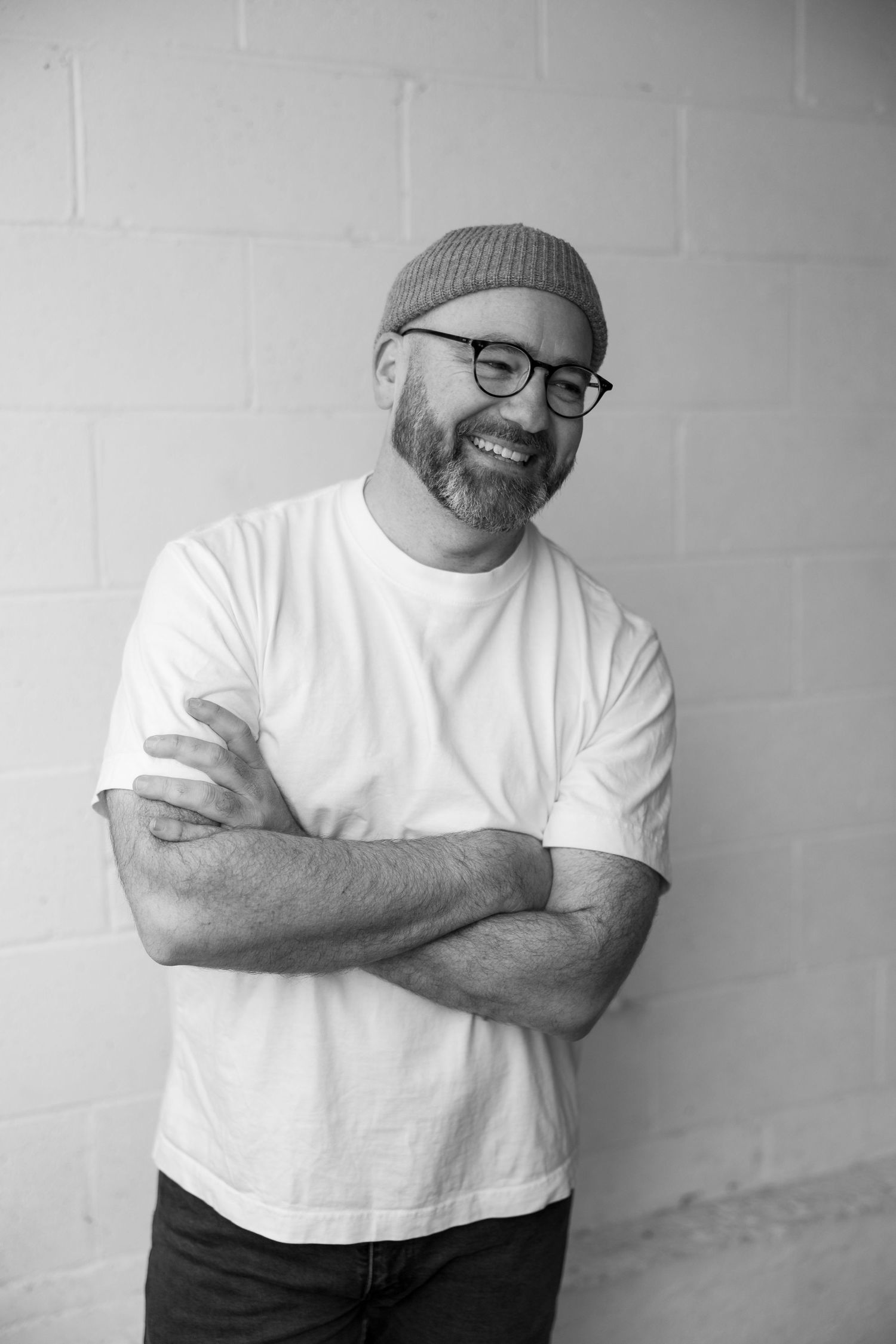 Black and white photo of a bearded man in glasses and beanie, smiling with arms crossed against a brick wall.