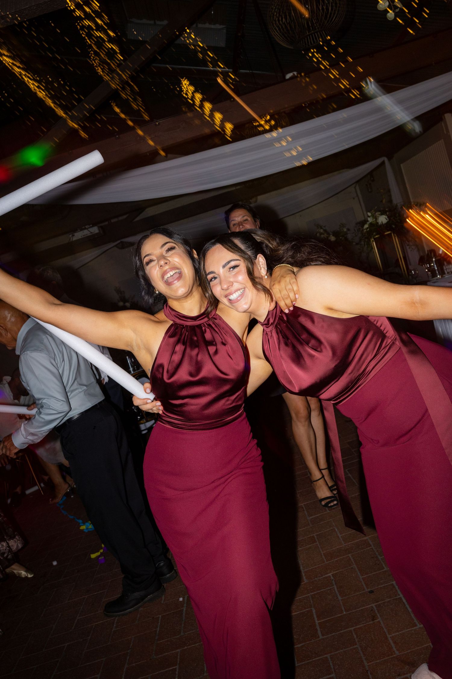 Two bridesmaids in burgundy satin halter gowns dancing joyfully at a wedding reception.