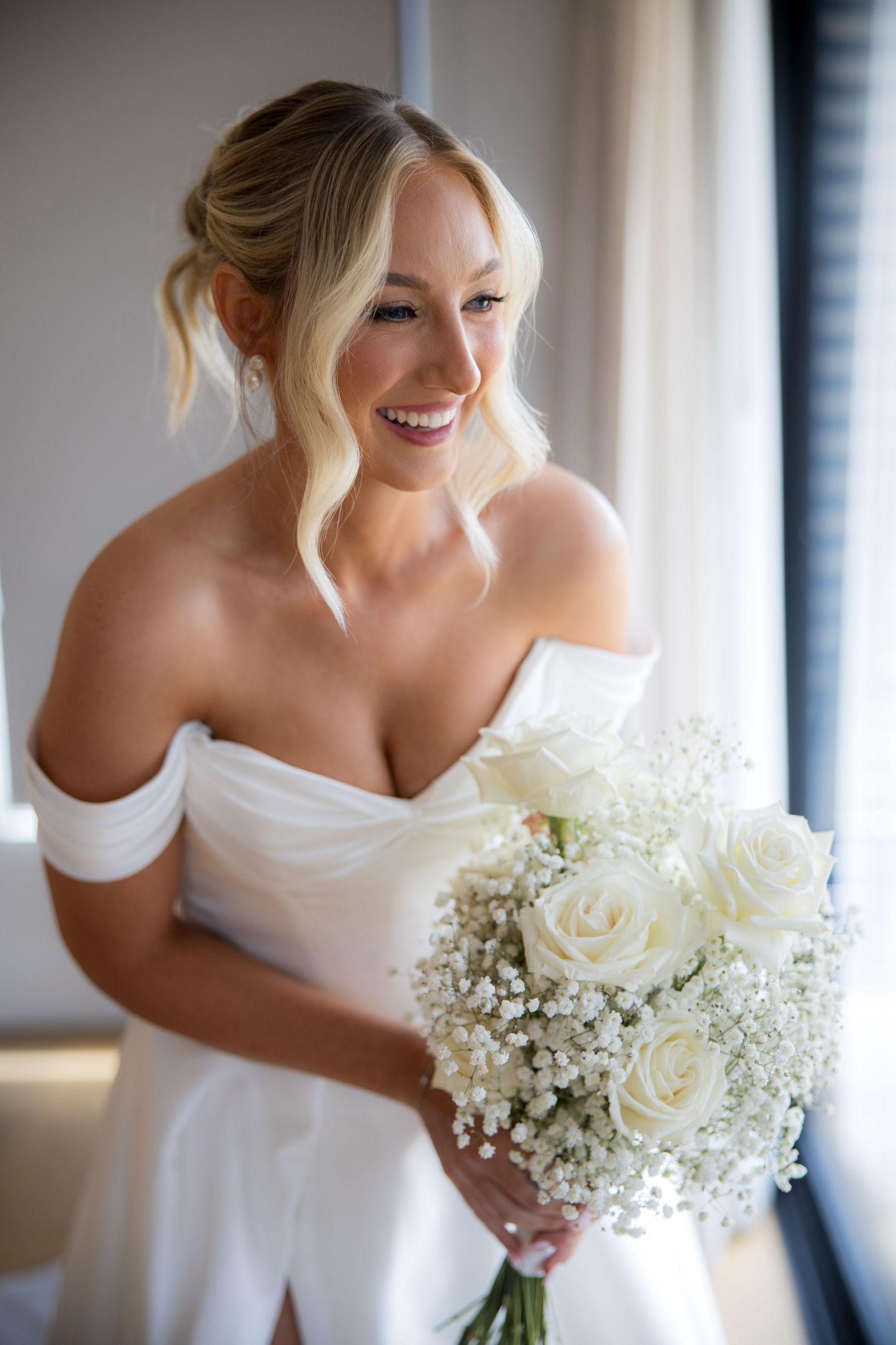 Radiant blonde bride in off-shoulder white gown holds bouquet of white roses and baby's breath indoors.