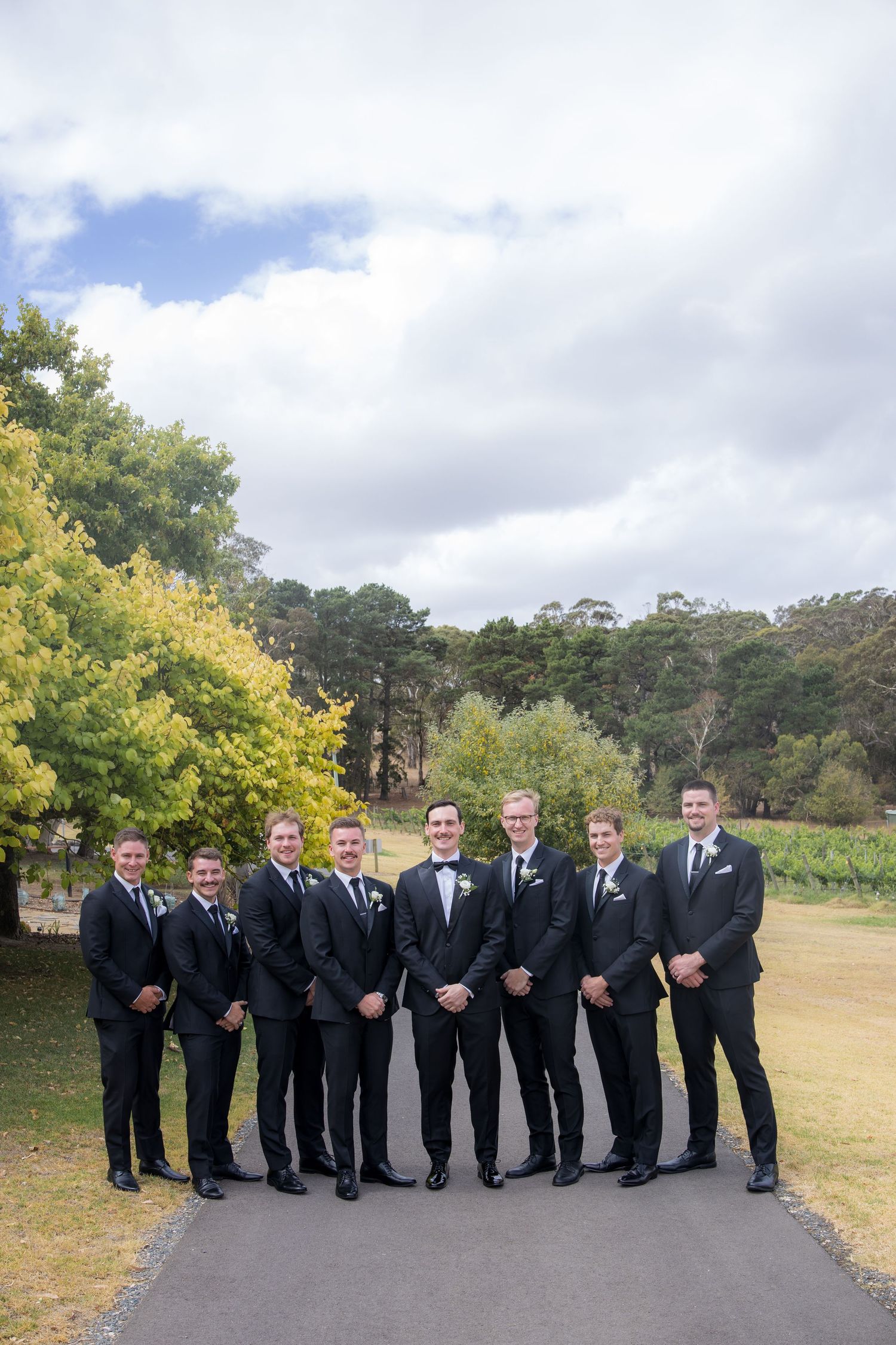 Groom and seven groomsmen in matching black suits posing on a pathway at outdoor venue.