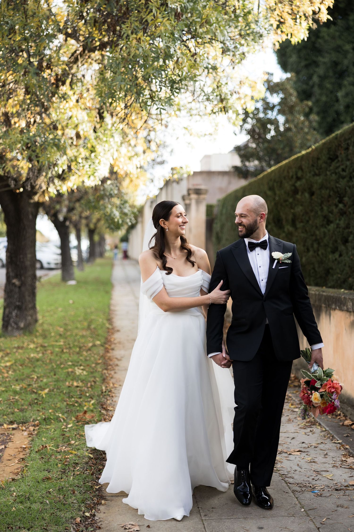 Bride and groom walking hand in hand on a tree-lined sidewalk, laughing together on their wedding day.