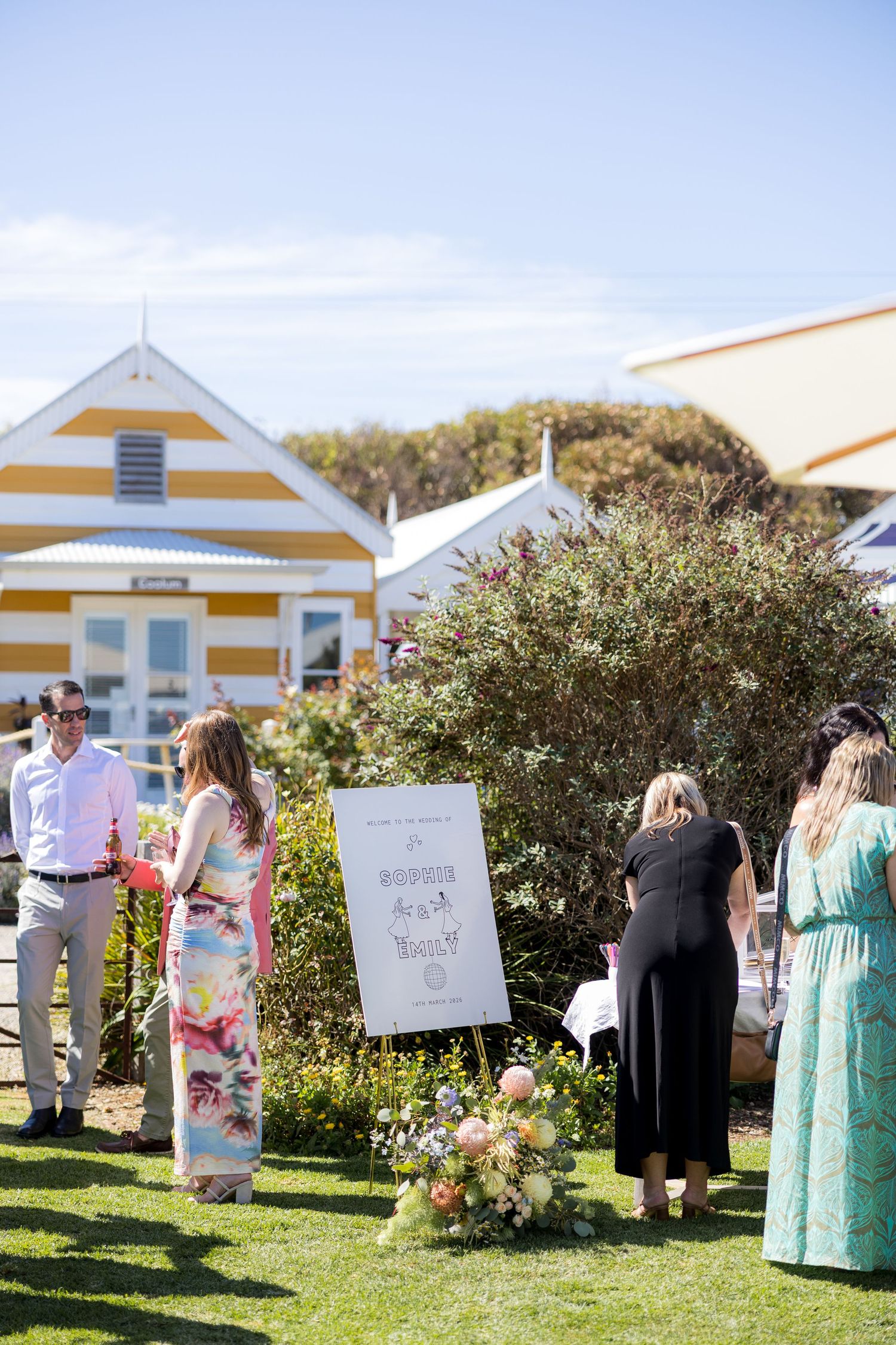 Wedding guests gather around a welcome sign on an easel at an outdoor garden ceremony.