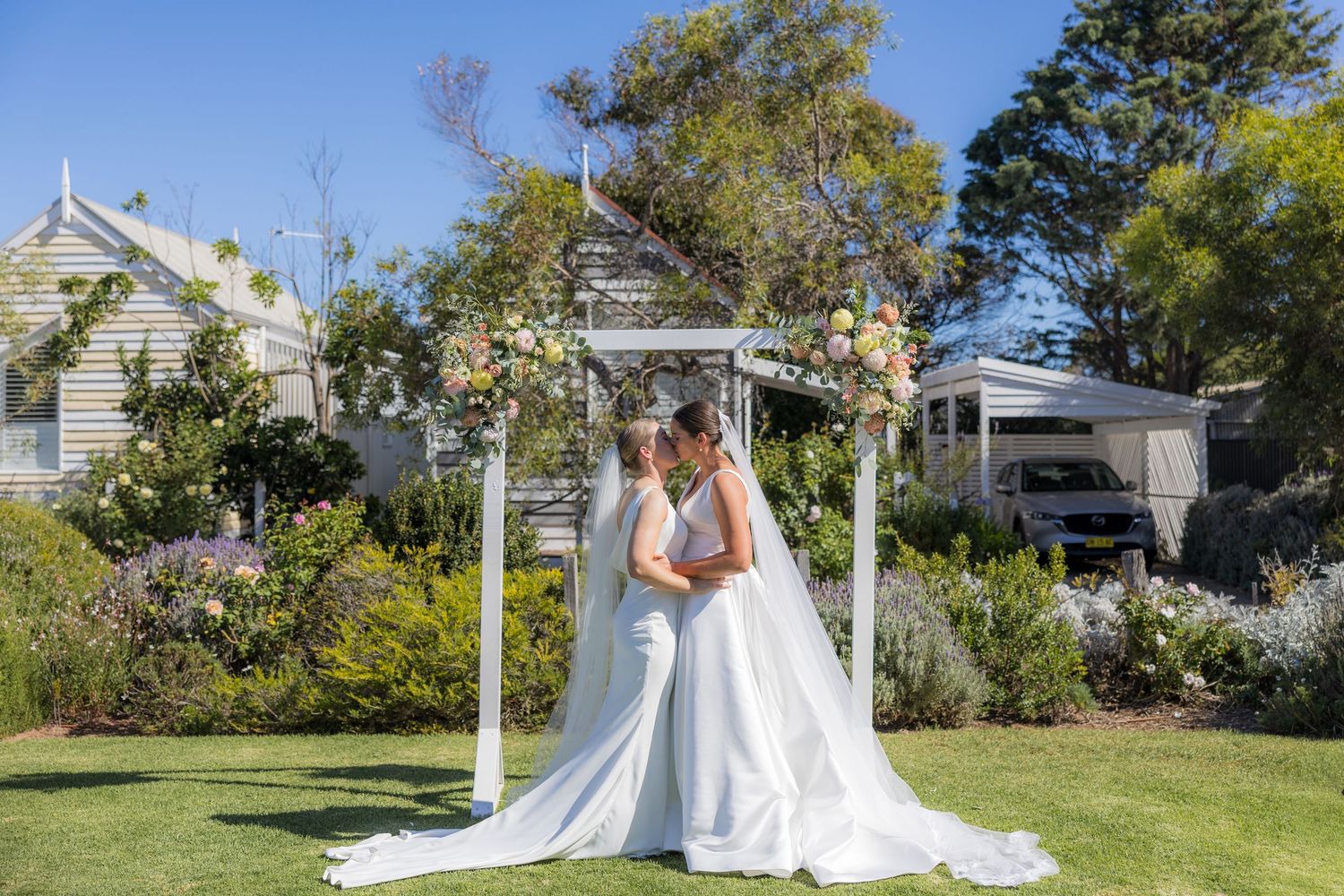 Two brides share a romantic kiss under a white floral arch in a sunny garden wedding setting.