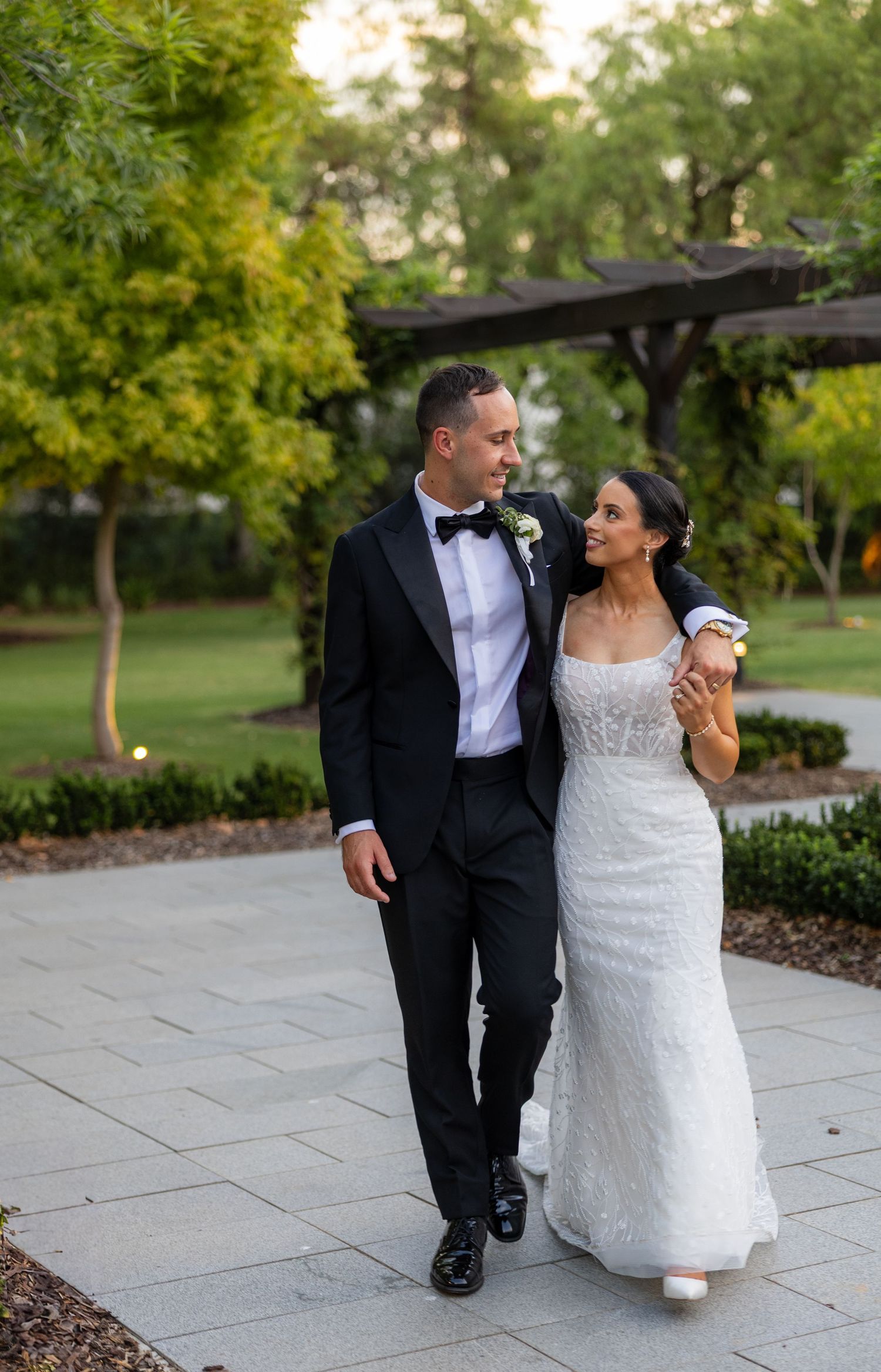 Newlyweds walk hand in hand along garden path, groom in tuxedo, bride in beaded gown.