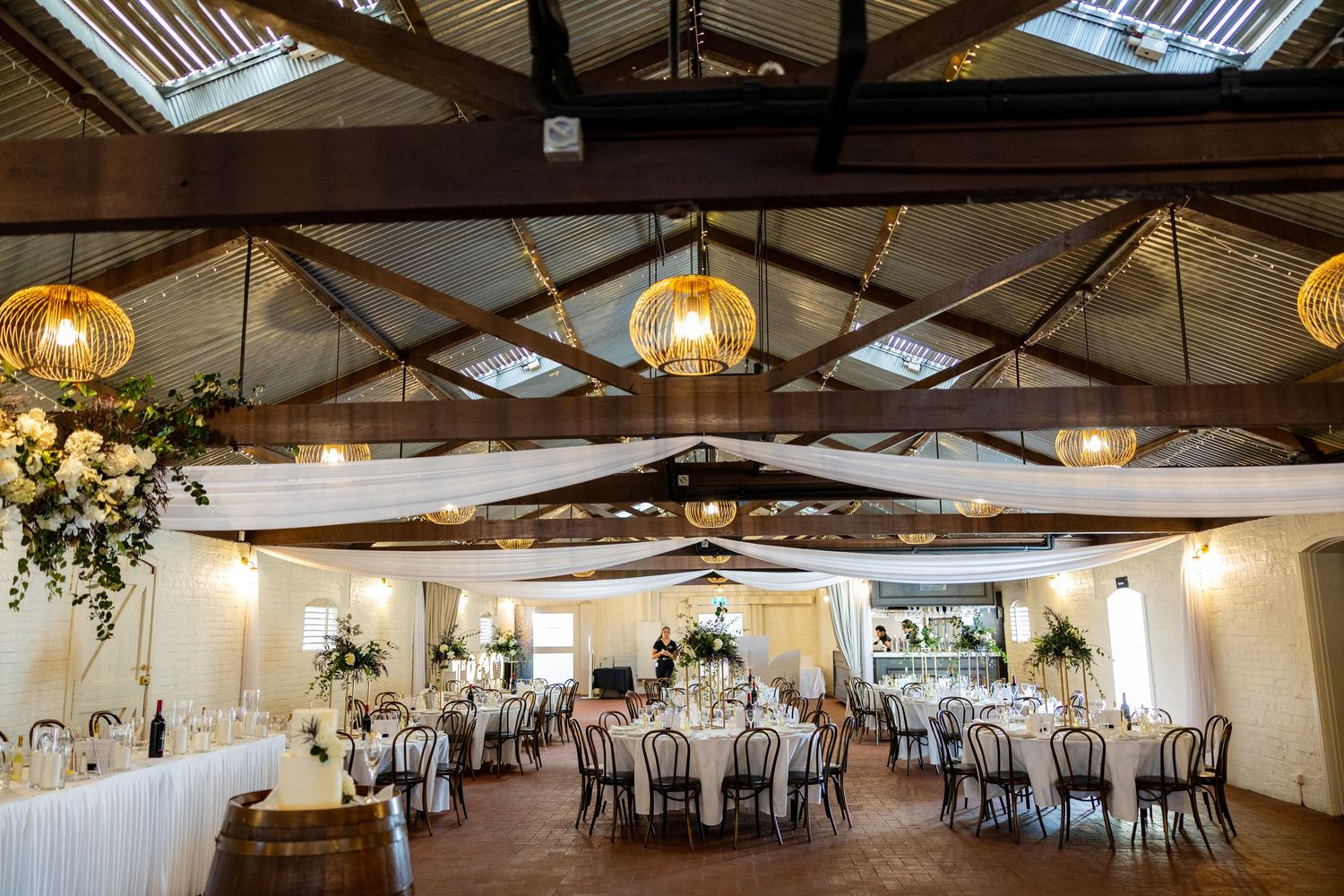 Rustic wedding reception hall with exposed timber roof, rattan pendant lights, and white floral decor.