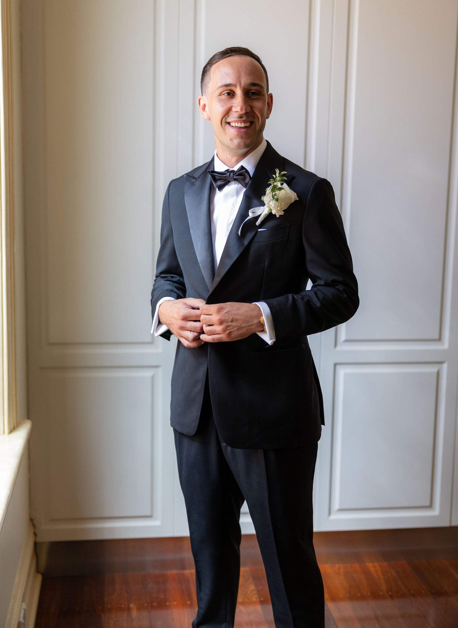 Smiling groom in black tuxedo with bow tie and boutonniere posing indoors.