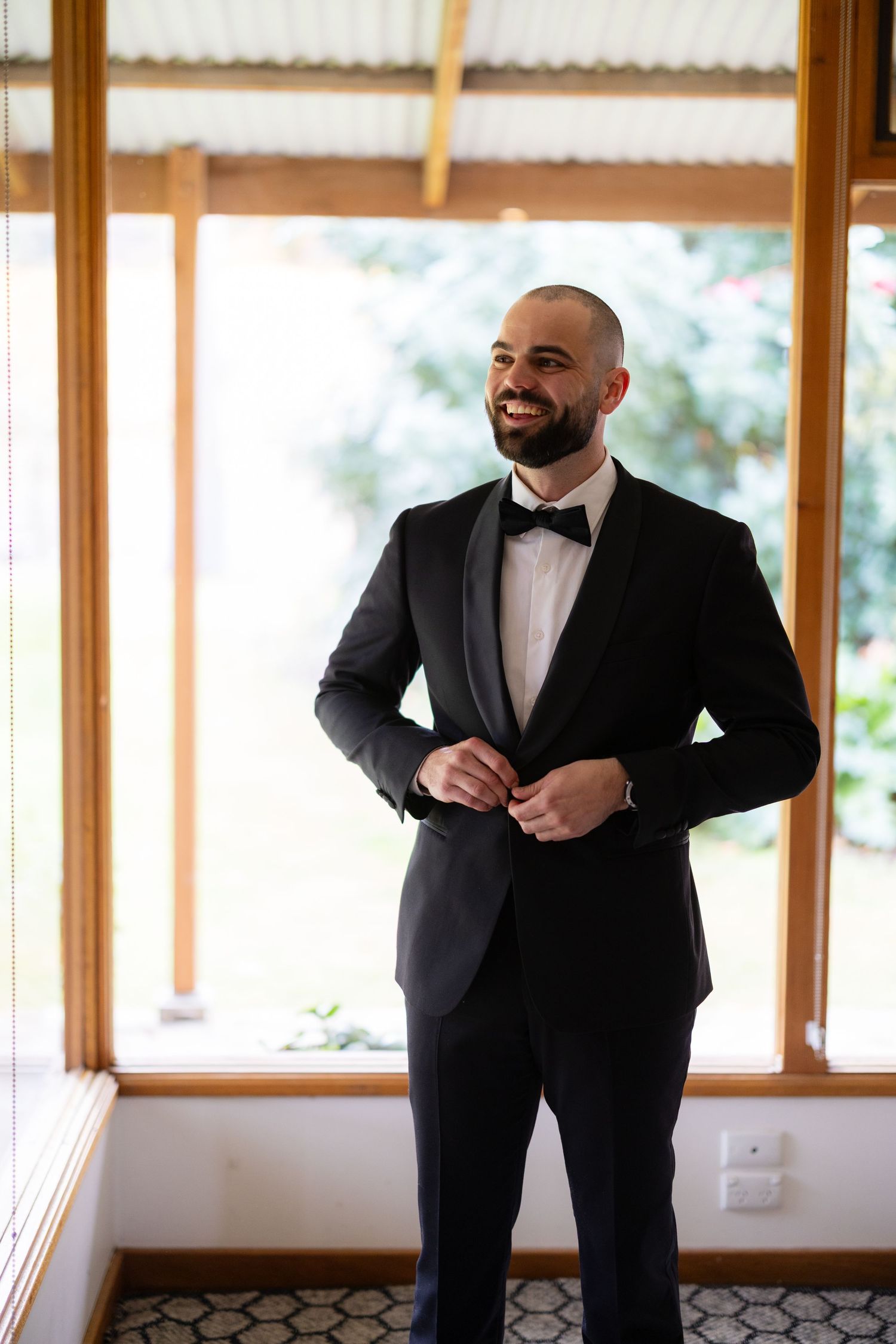 Handsome groom in black tuxedo with bow tie smiling while buttoning jacket near window.