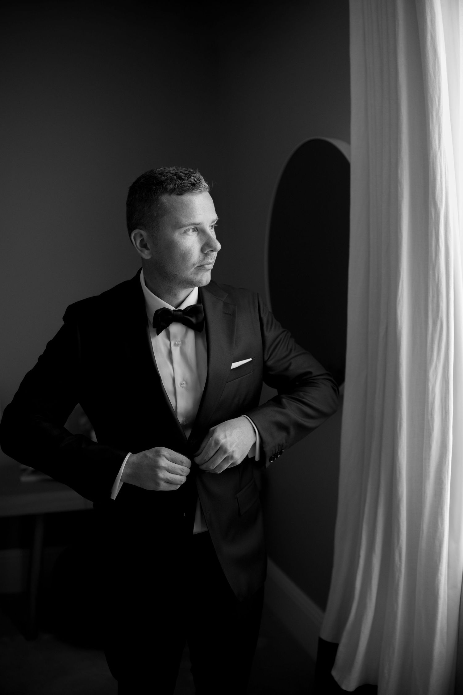 Elegant groom in black tuxedo and bow tie gazes out window in black and white portrait.