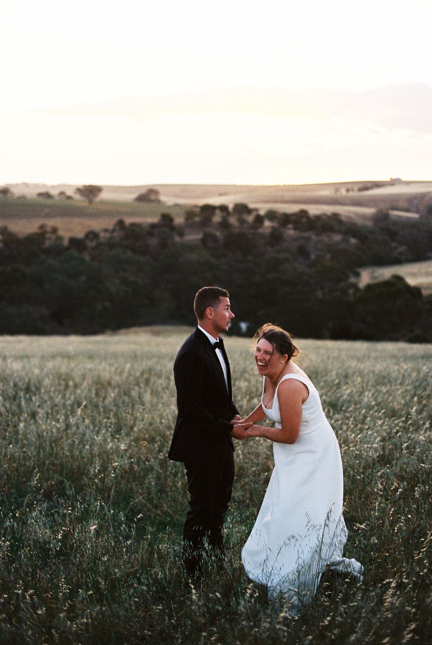 Bride and groom laughing together in a golden field with scenic countryside at sunset.