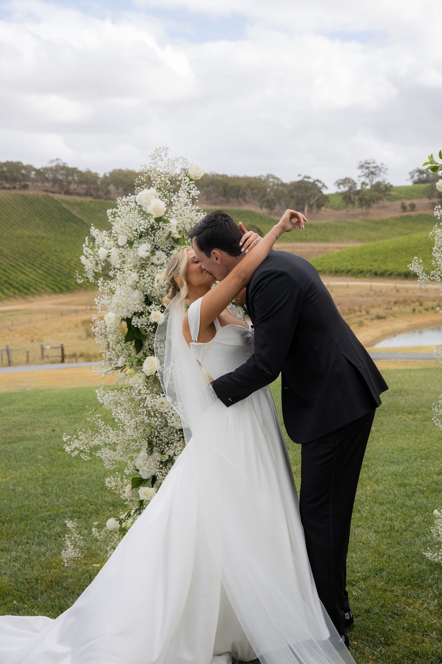 Bride and groom share first kiss at outdoor vineyard wedding ceremony with white floral arch.