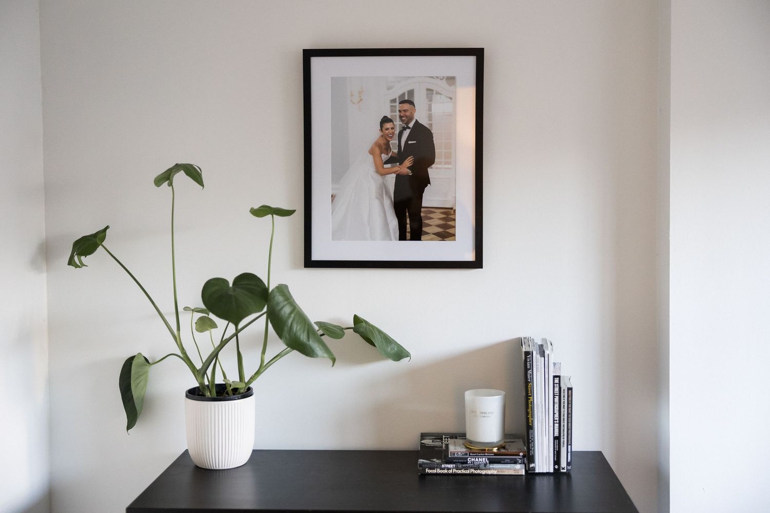Wedding portrait in black frame above dark dresser with monstera plant and decorative books.