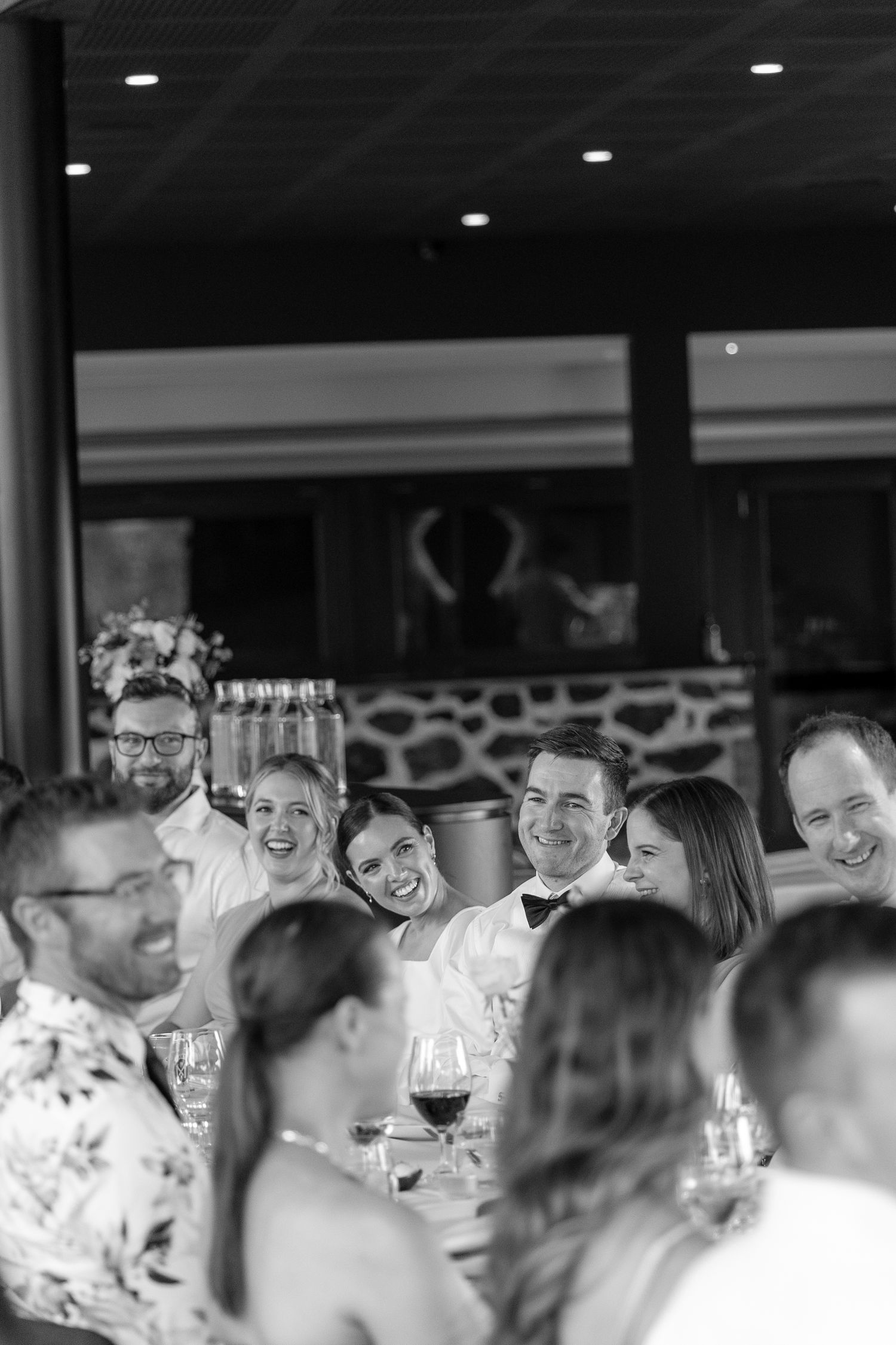 Black and white wedding reception photo of smiling bride, groom and guests seated at dinner table.