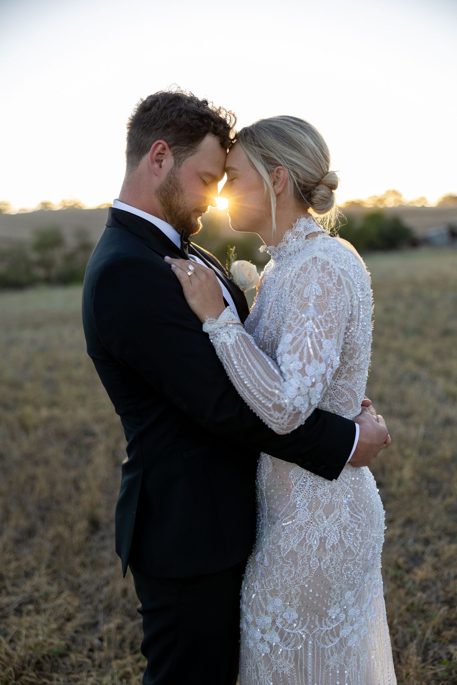 Bride and groom embrace forehead-to-forehead in a sun-kissed field at golden hour sunset.