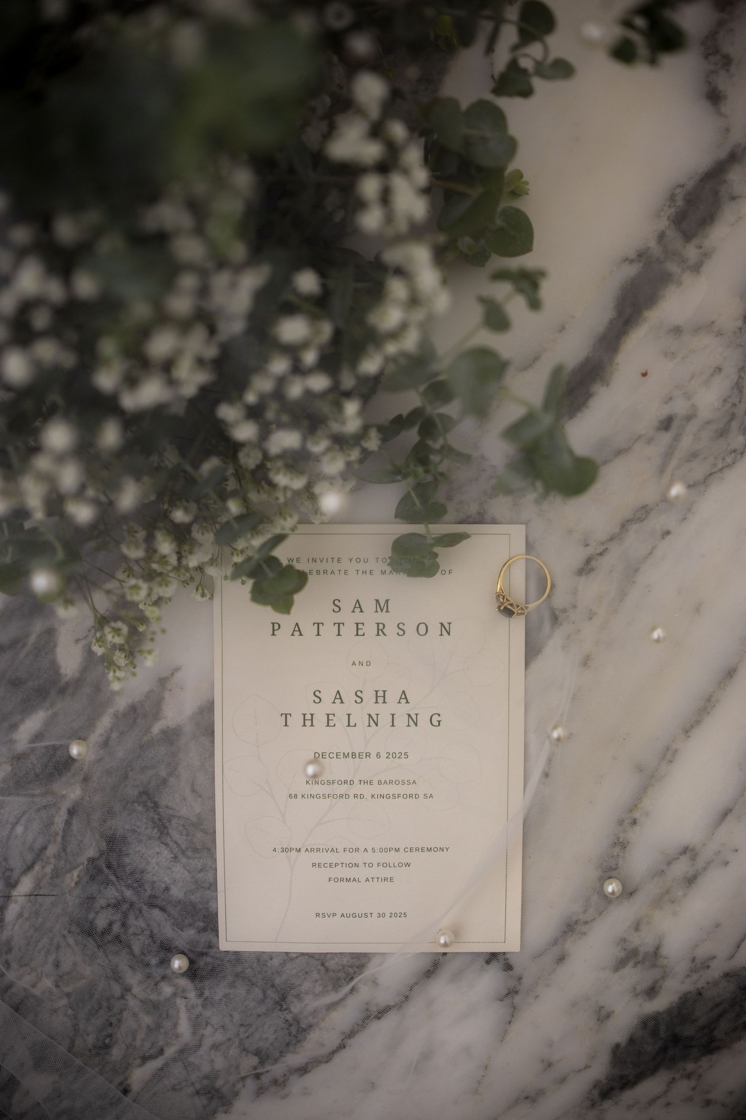Wedding invitation for Sam Patterson and Sasha Thelning on marble surface with flowers and ring.