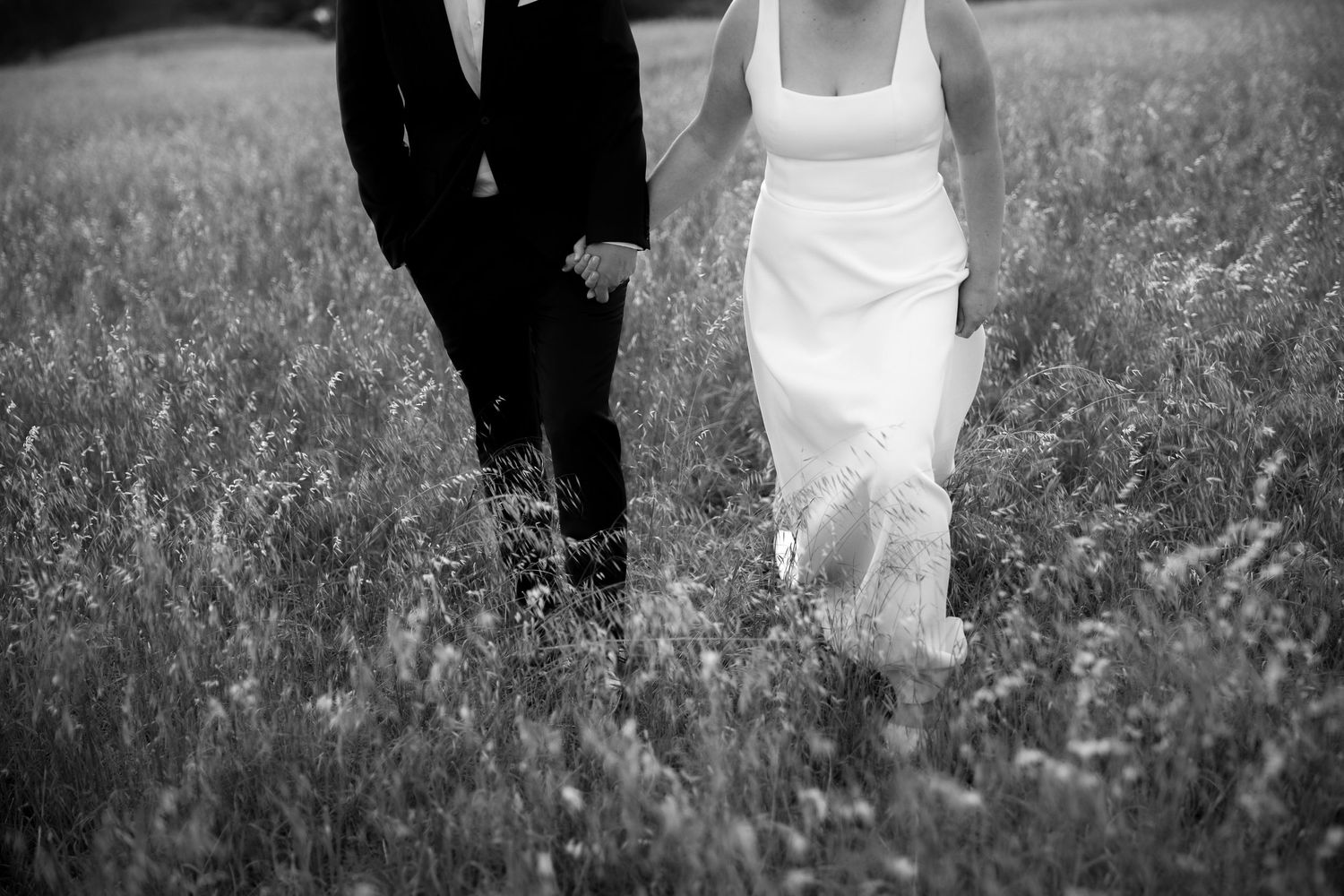 Black and white photo of bride and groom holding hands walking through a wildflower meadow.