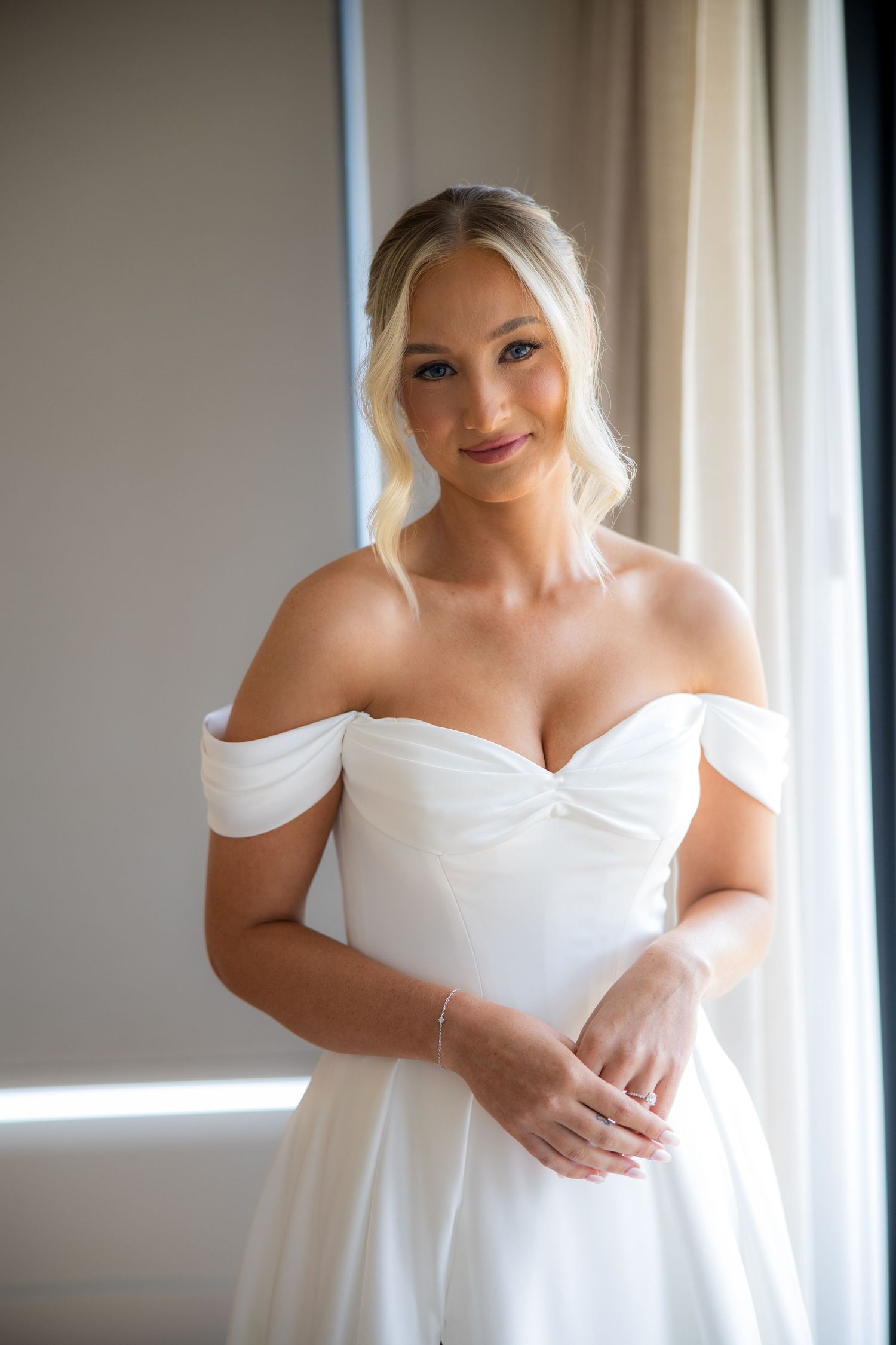 Bride in off-shoulder white wedding gown posing elegantly near a window on her wedding day.
