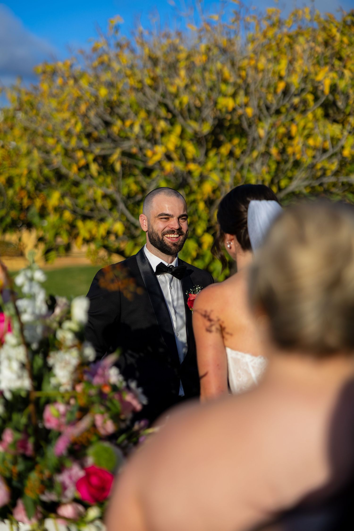 Smiling groom in black tuxedo looking at bride during outdoor wedding ceremony with colorful floral arrangements.