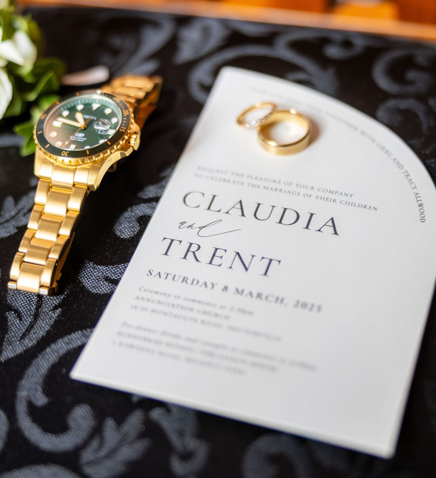 Wedding invitation for Claudia and Trent, March 8 2025, beside gold Rolex watch and rings.