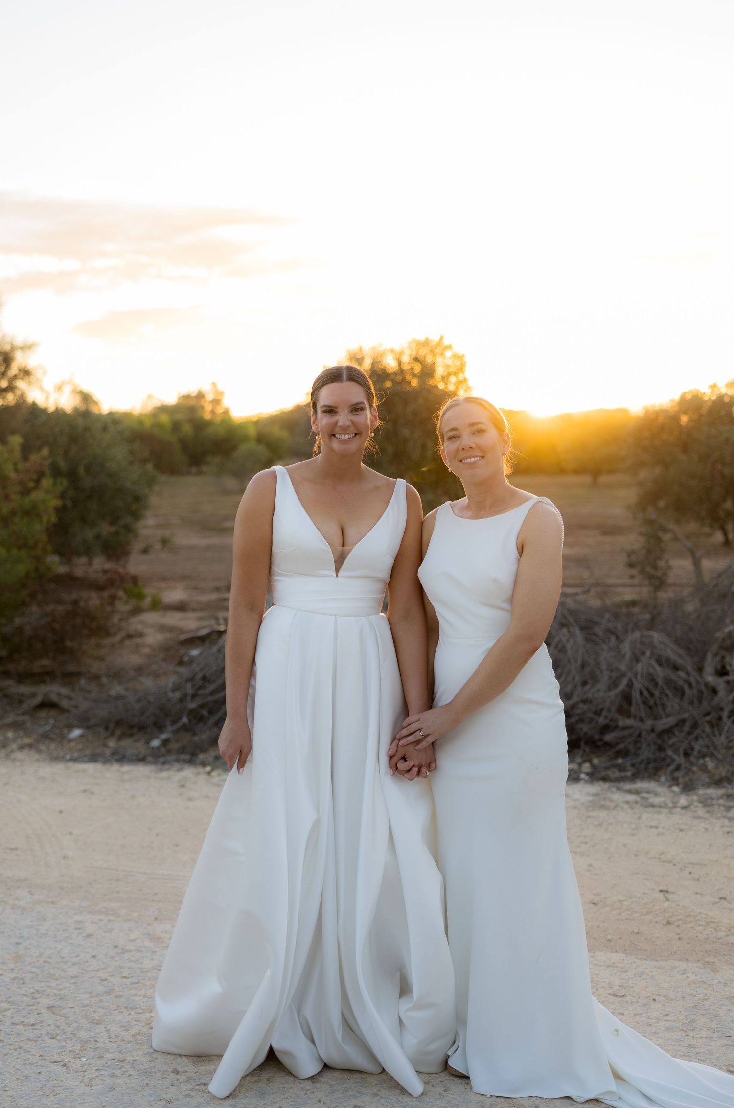 Two brides in white gowns smile hand-in-hand during a golden sunset outdoor wedding portrait.