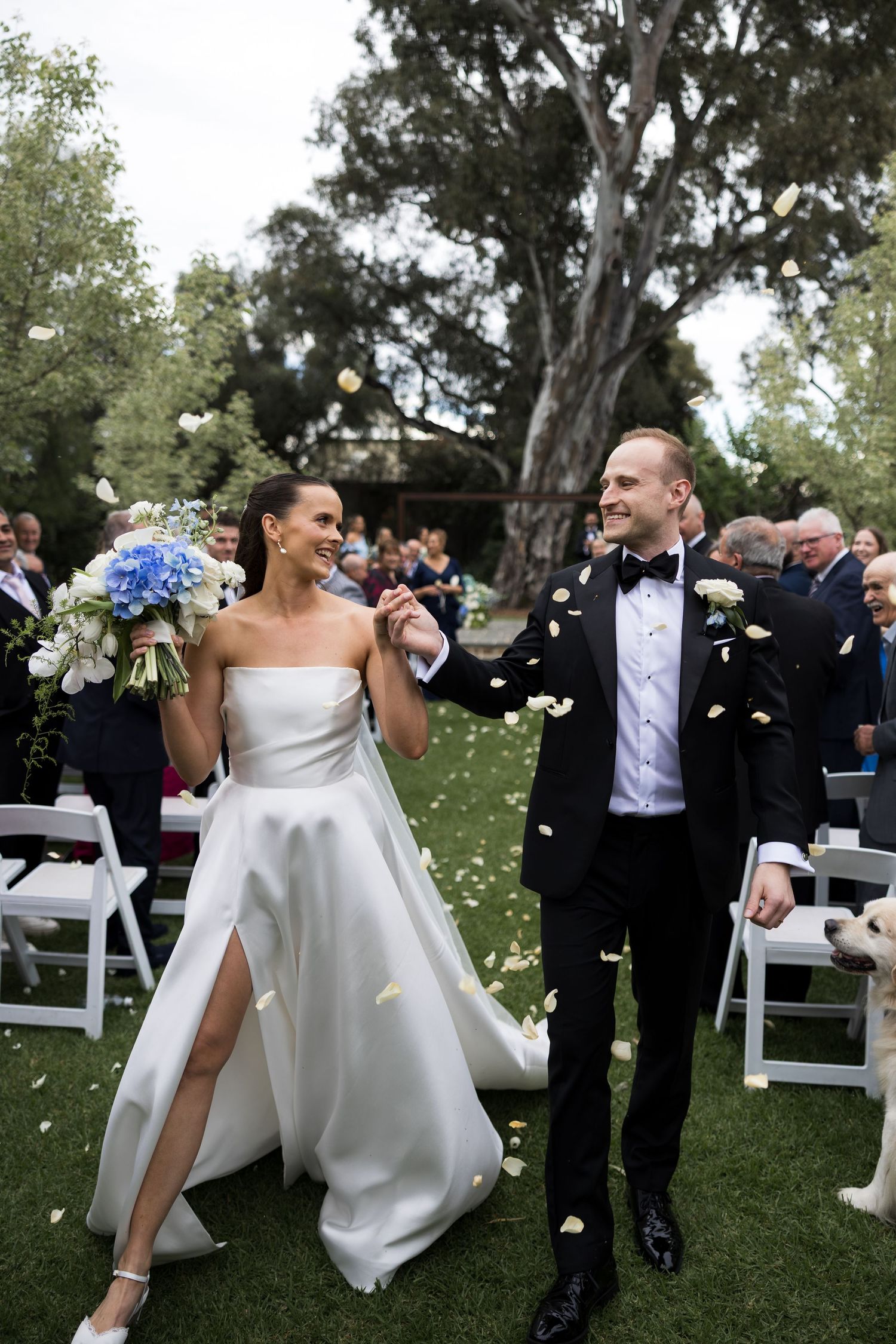 Smiling bride in a strapless satin gown holding a blue bouquet walks hand in hand with a groom in a tuxedo as petals fall outdoors.