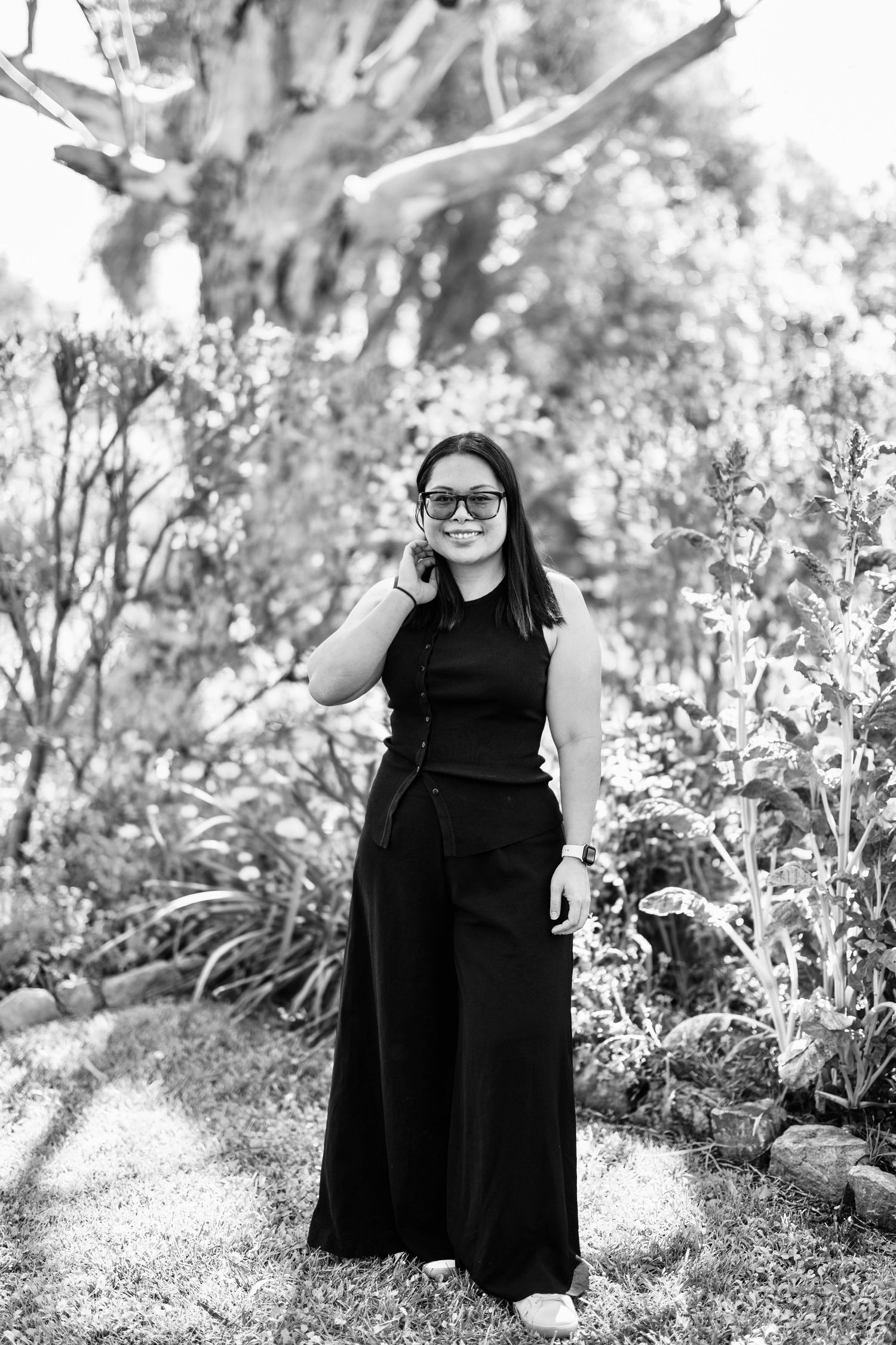 Black and white portrait of a young woman in sunglasses and black jumpsuit standing in a lush garden.