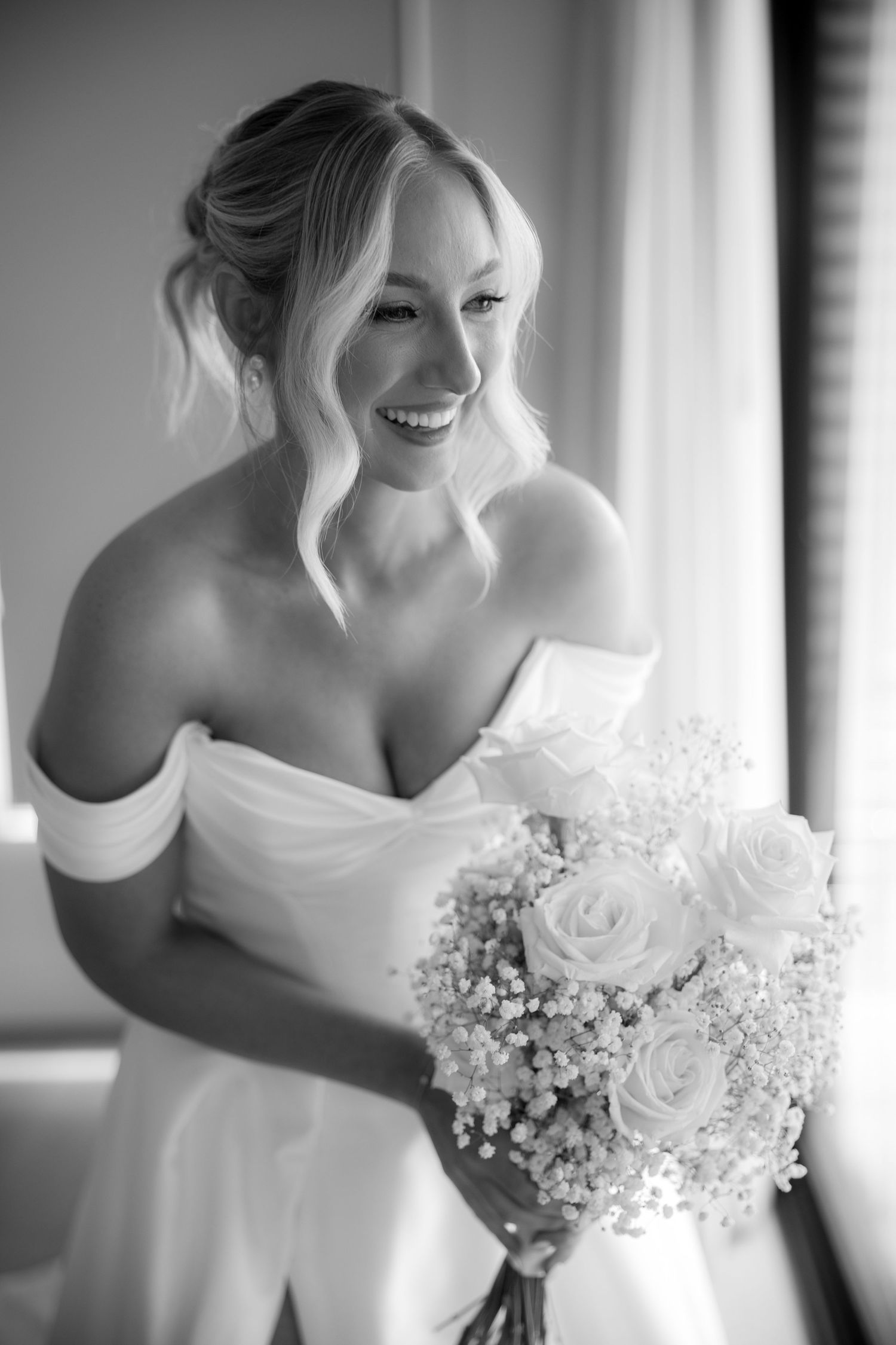 Black and white portrait of a laughing bride holding a bouquet in an off-shoulder wedding gown.